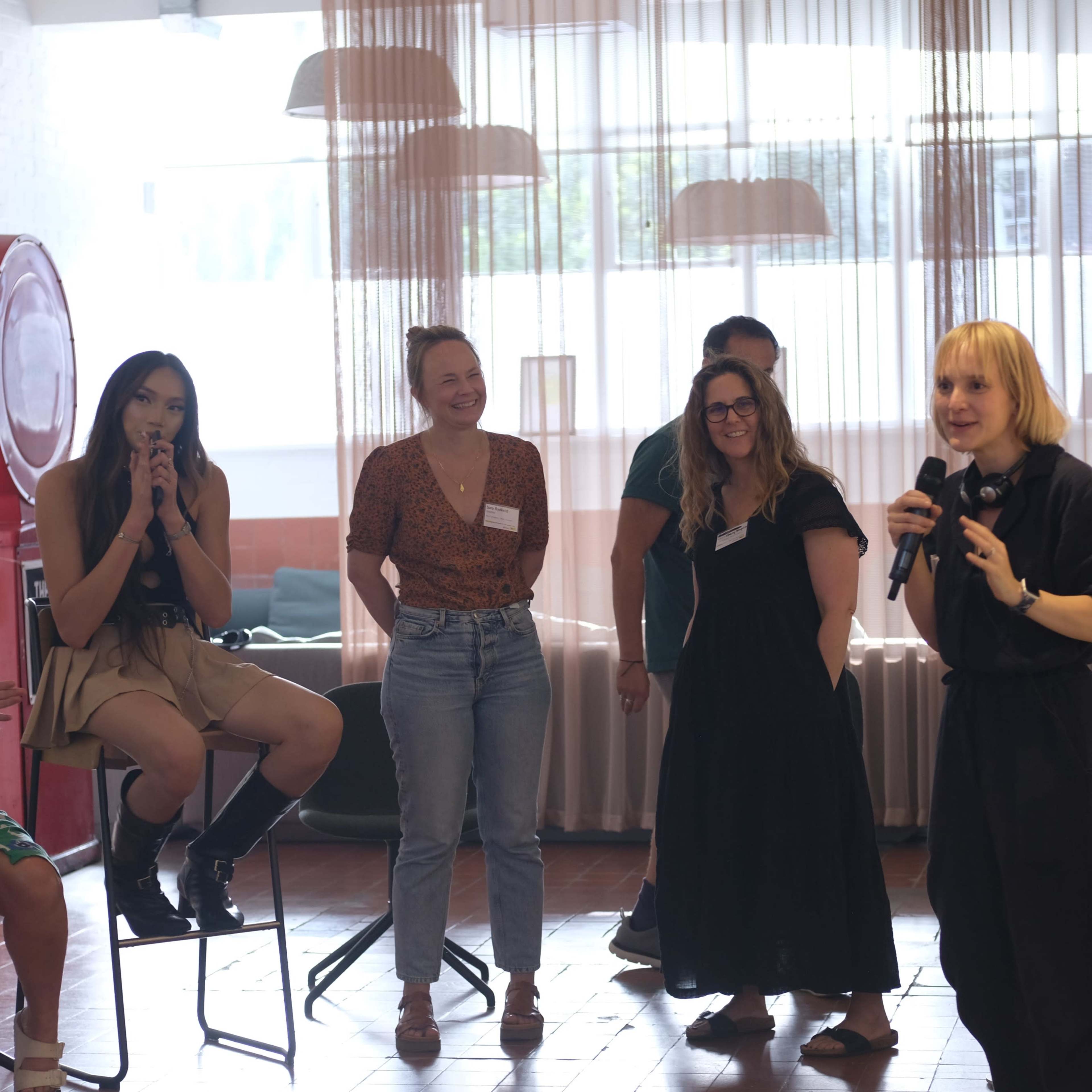 A group of women engages in a discussion while one speaker addresses the audience using a microphone in a brightly lit room.