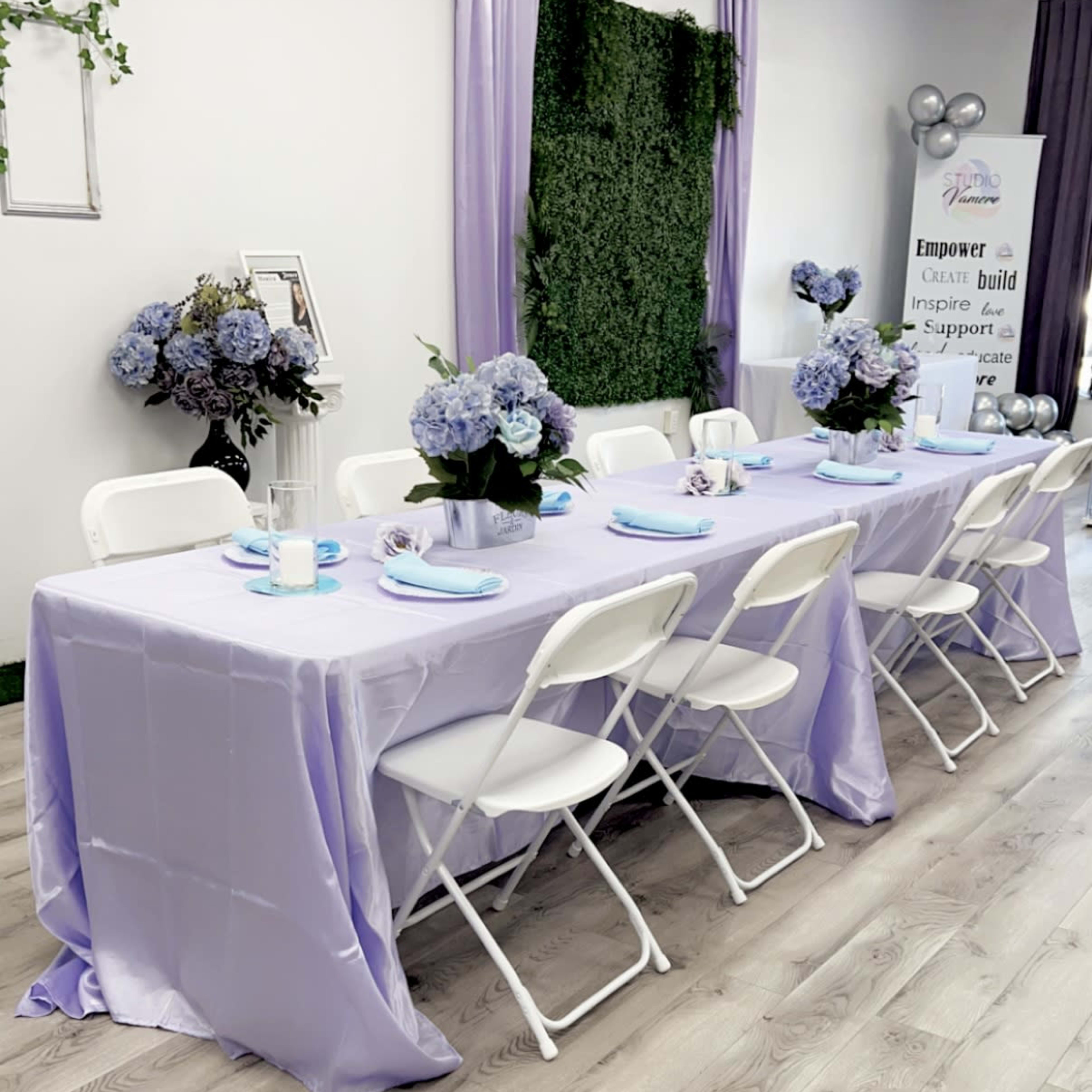 A table set for an event, covered with a light purple tablecloth and surrounded by white chairs, features decorative floral arrangements and a banner promoting empowerment and support.