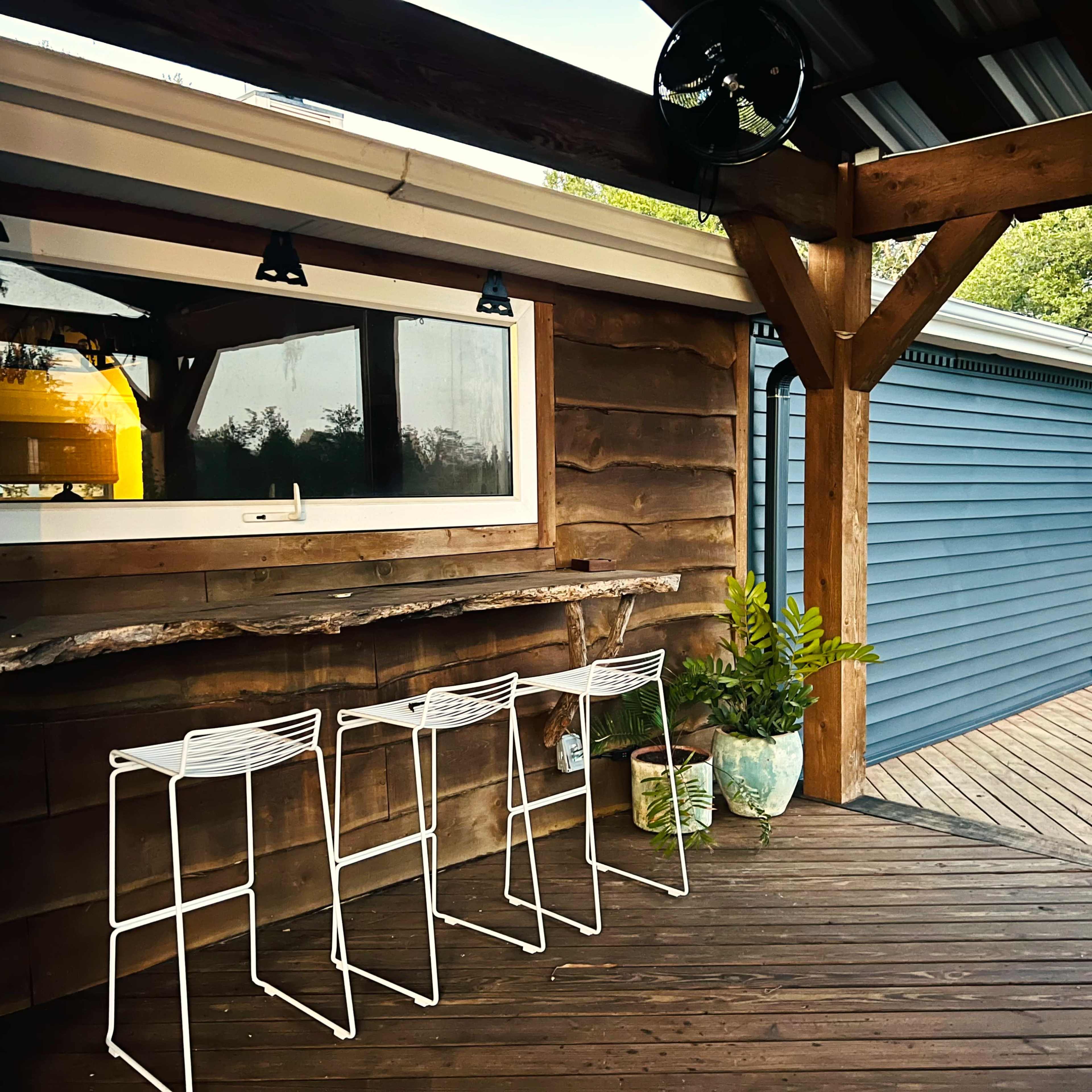 A wooden deck with three white metal bar stools positioned in front of a rustic wooden counter, beside a blue-painted wall of a house.