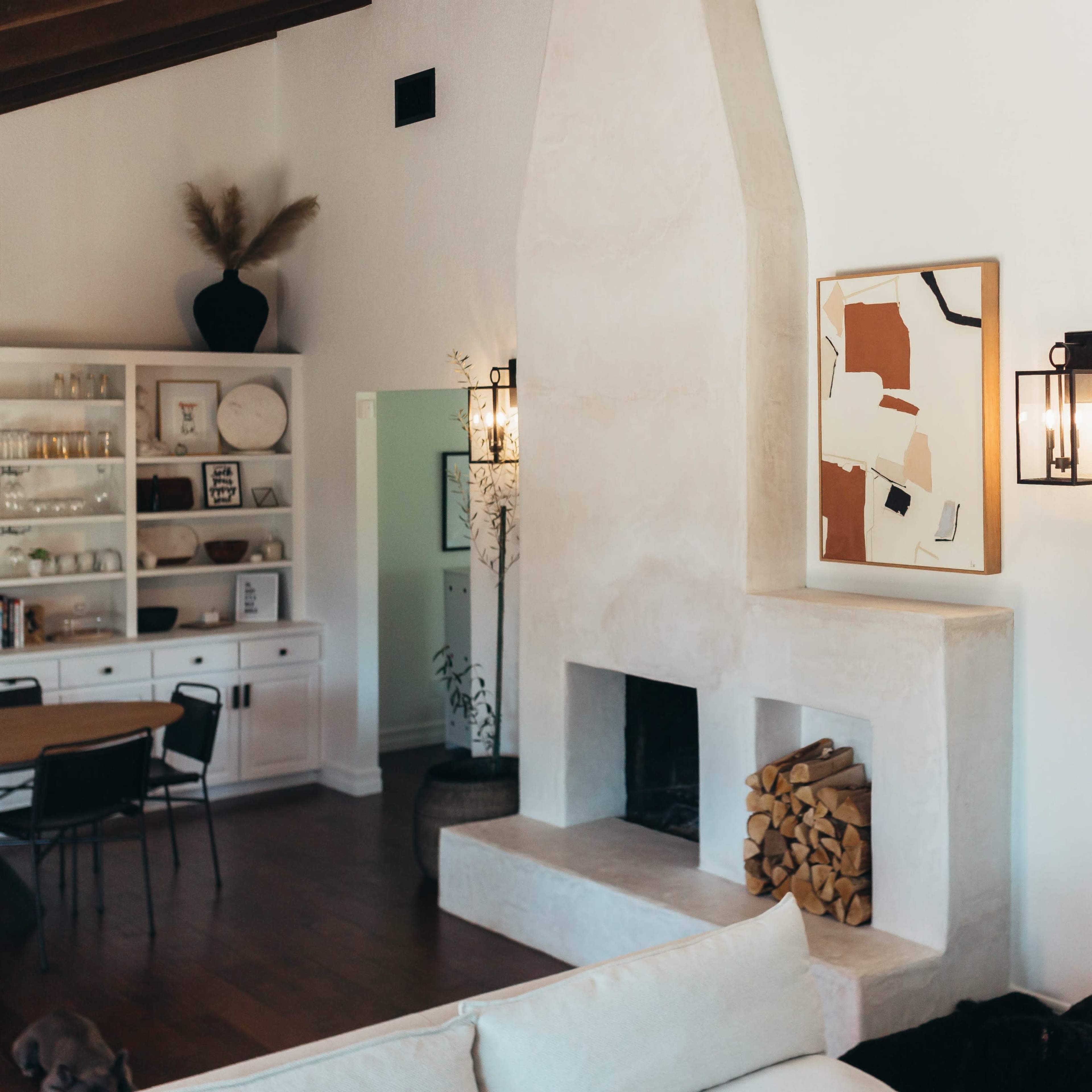 A modern living room featuring a white plaster fireplace, a piece of artwork on the wall, and decorative mirrors beside a stacked pile of firewood.
