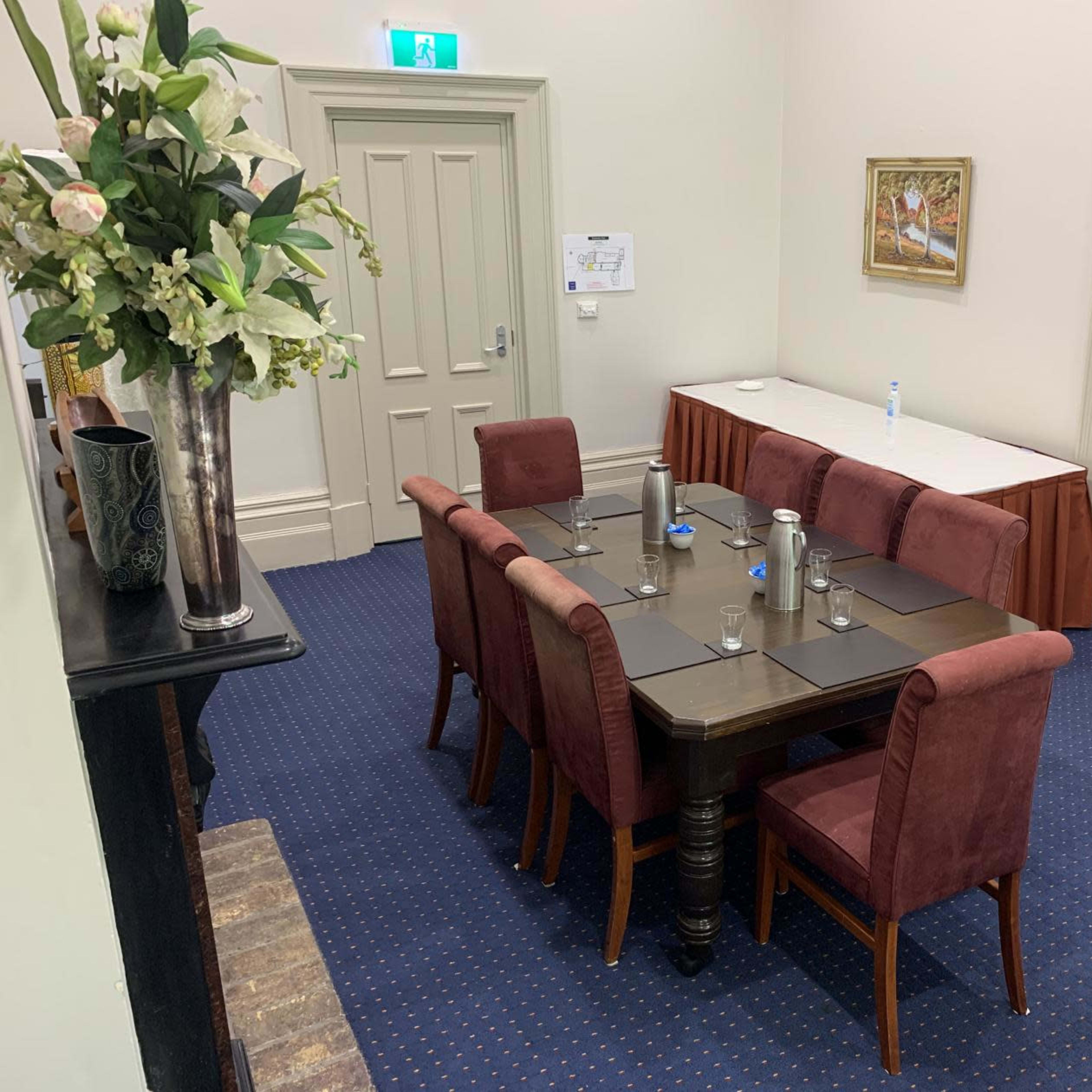A rectangular table with eight upholstered chairs is set in a meeting room, accompanied by a small side table and a flower arrangement.