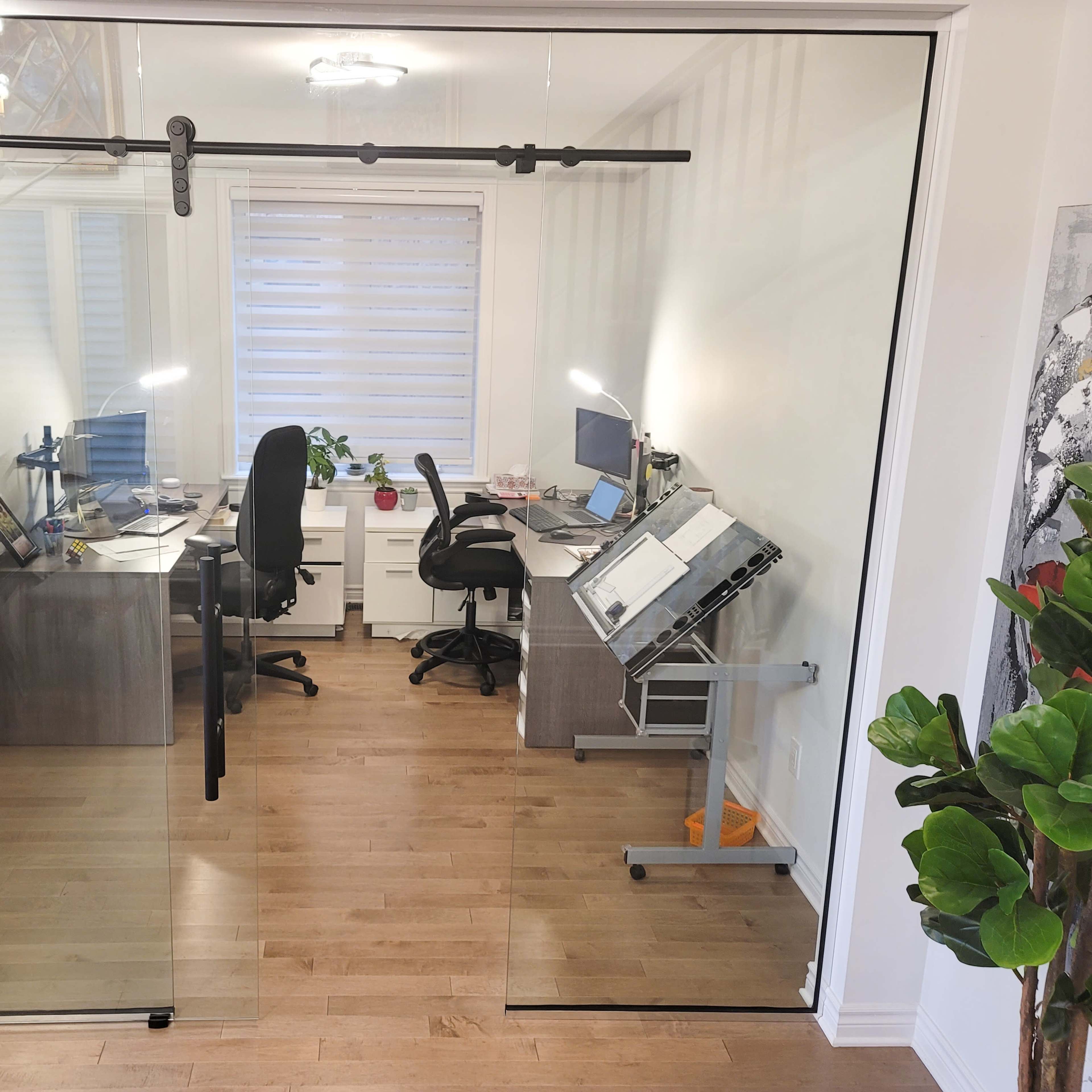 A modern office space divided by a sliding glass door, featuring two desks with computers and an art easel in a well-lit environment.