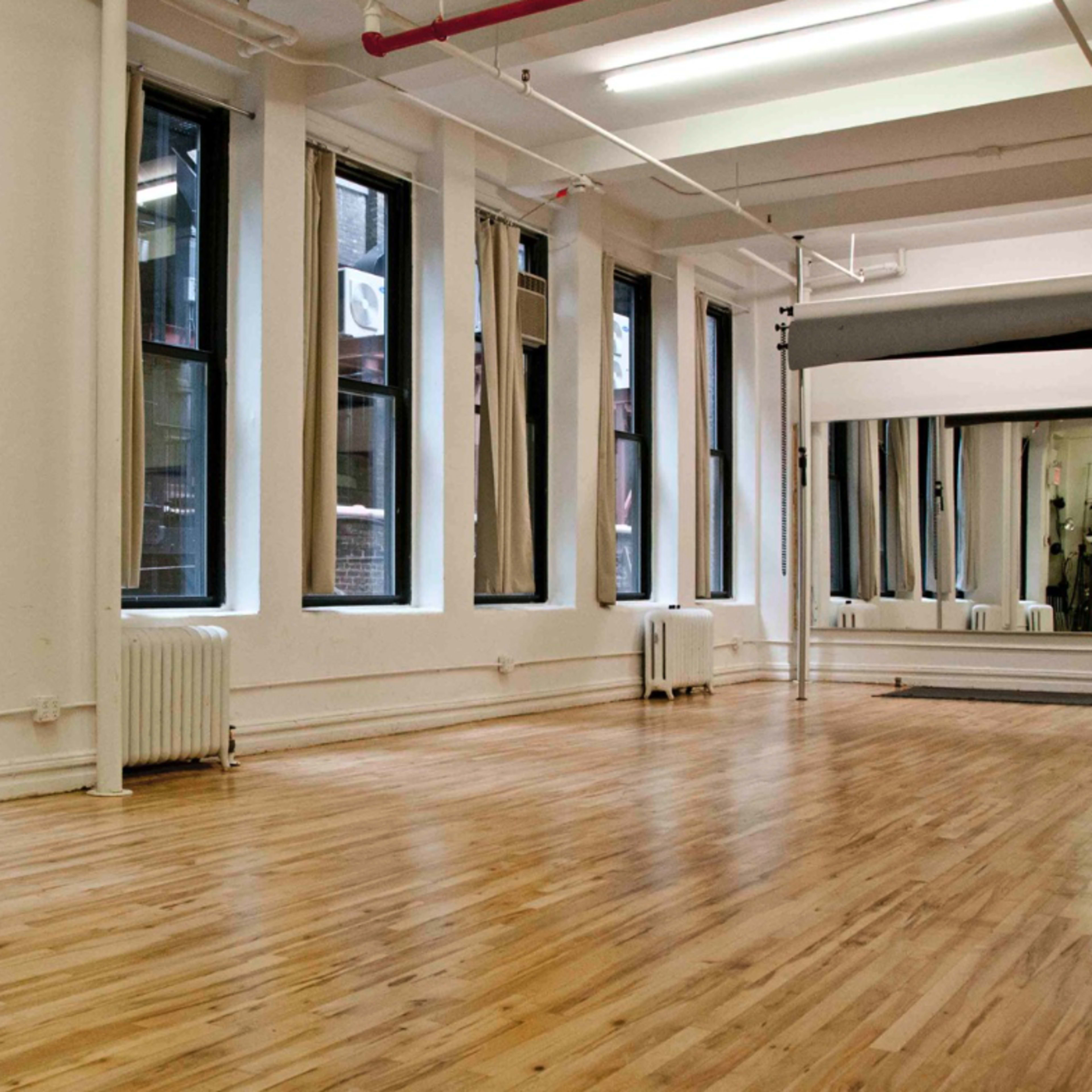 A spacious, empty studio with large windows, wooden flooring, and minimal furnishings.