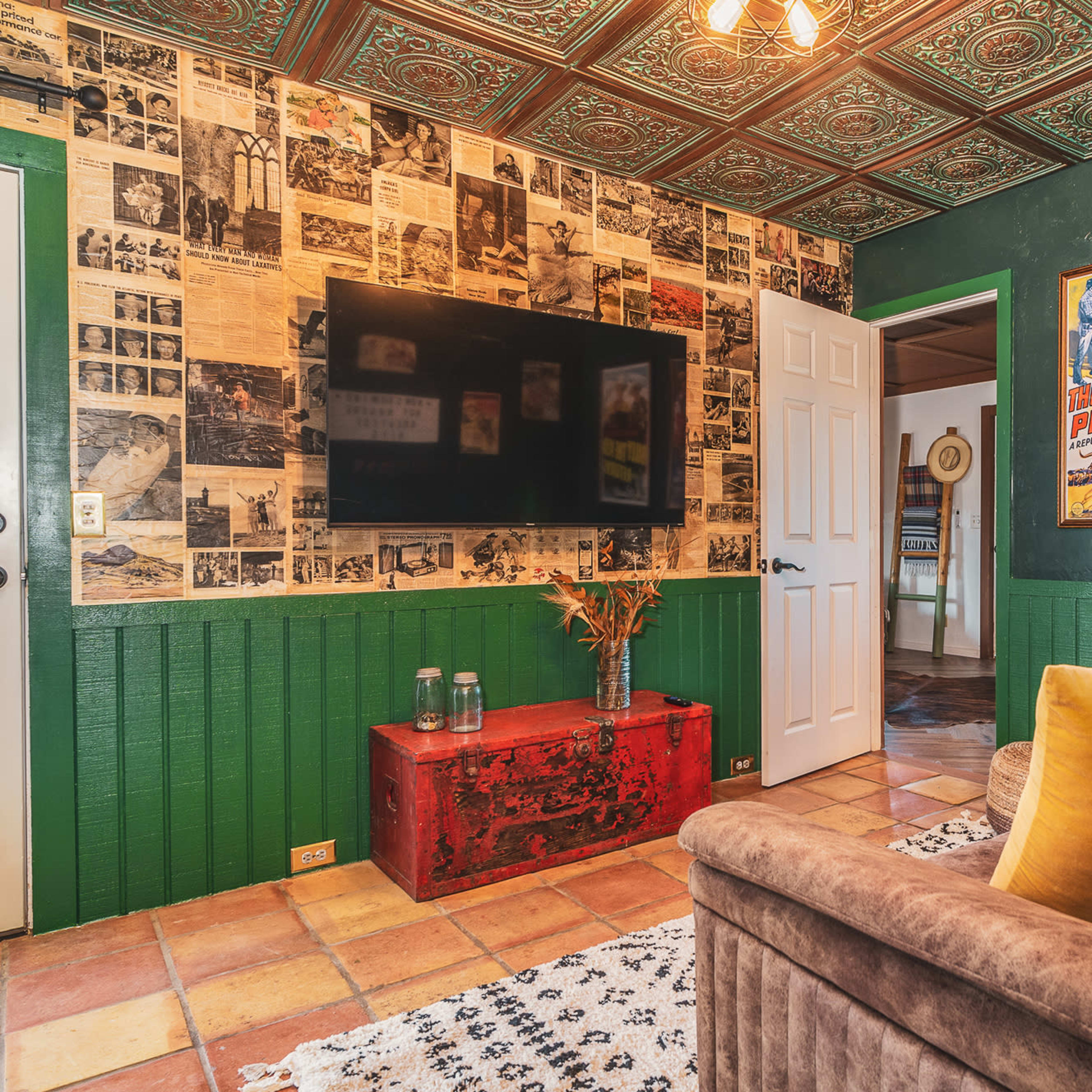 The room features a wall adorned with vintage newspaper clippings and posters, a television mounted on the wall, and a cozy seating area with a patterned rug.