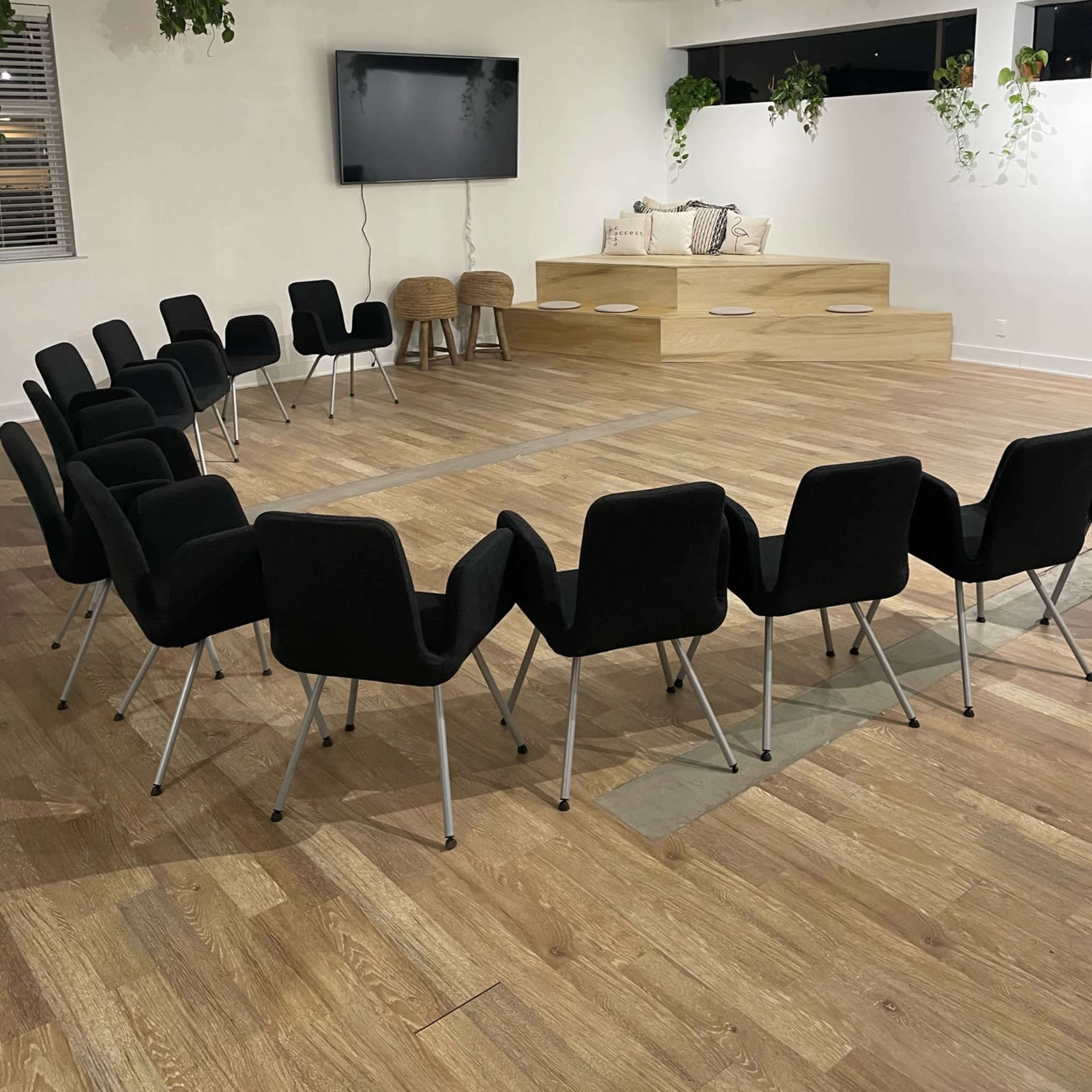 A circular arrangement of black chairs is set up in a brightly lit room with a large screen and a seating area against the wall.