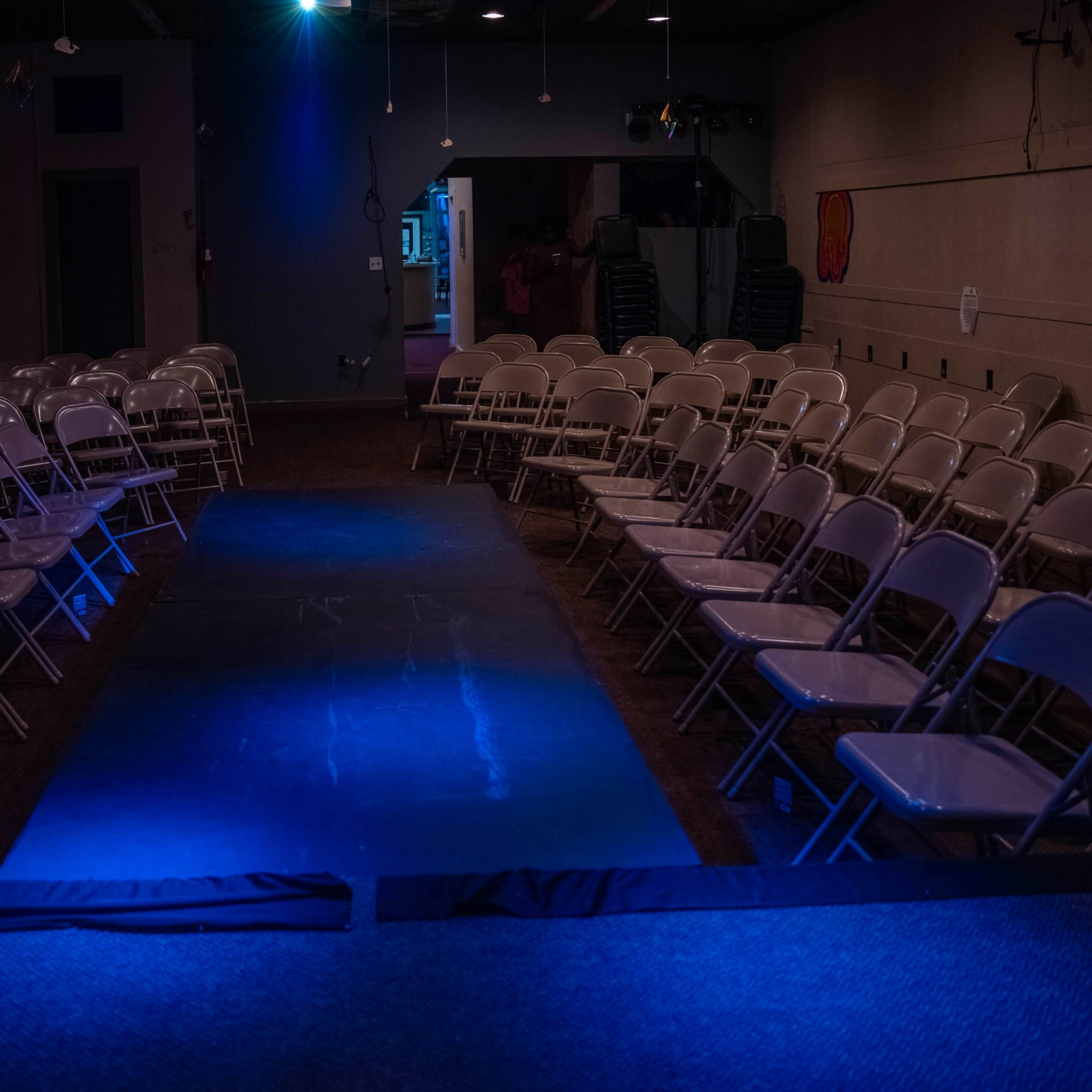 A dimly lit indoor venue features rows of white folding chairs facing a central runway illuminated by a blue light.