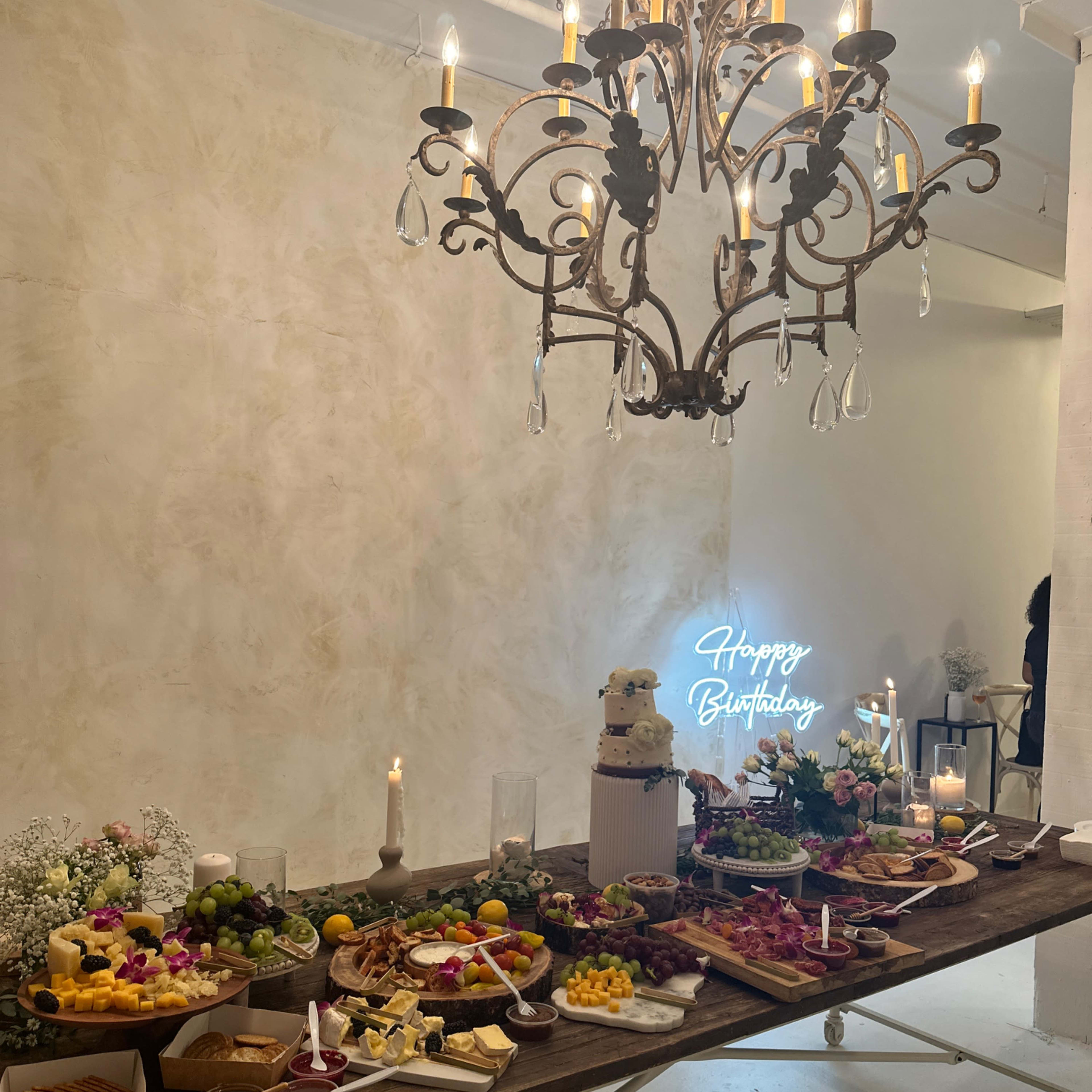 A large table is elegantly arranged with a variety of foods, including fruits, cheeses, and meats, under a chandelier, with a neon sign that says "Happy Birthday" in the background.