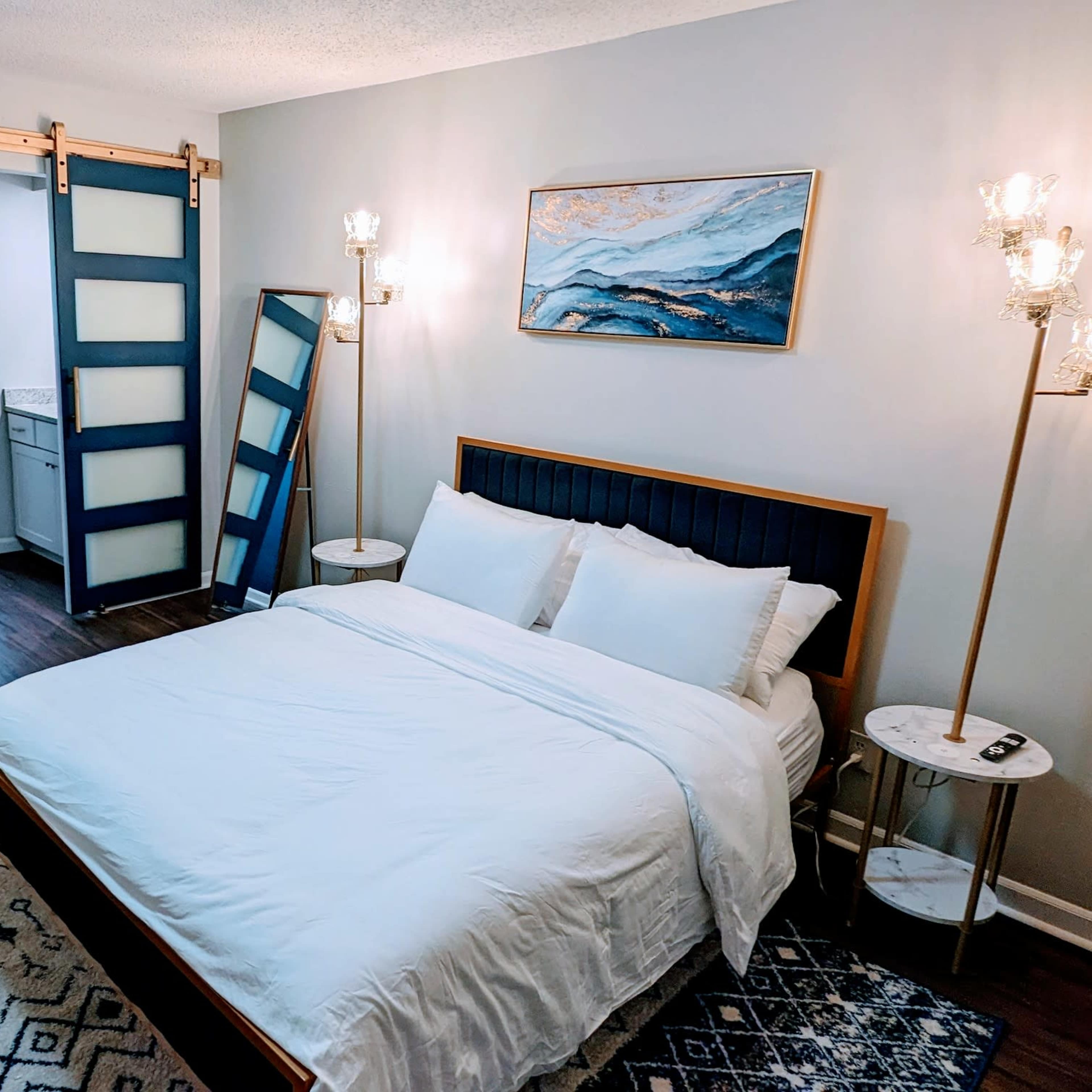 A well-lit bedroom features a bed with white linens, a large mirror, a glass sliding door, and modern light fixtures.