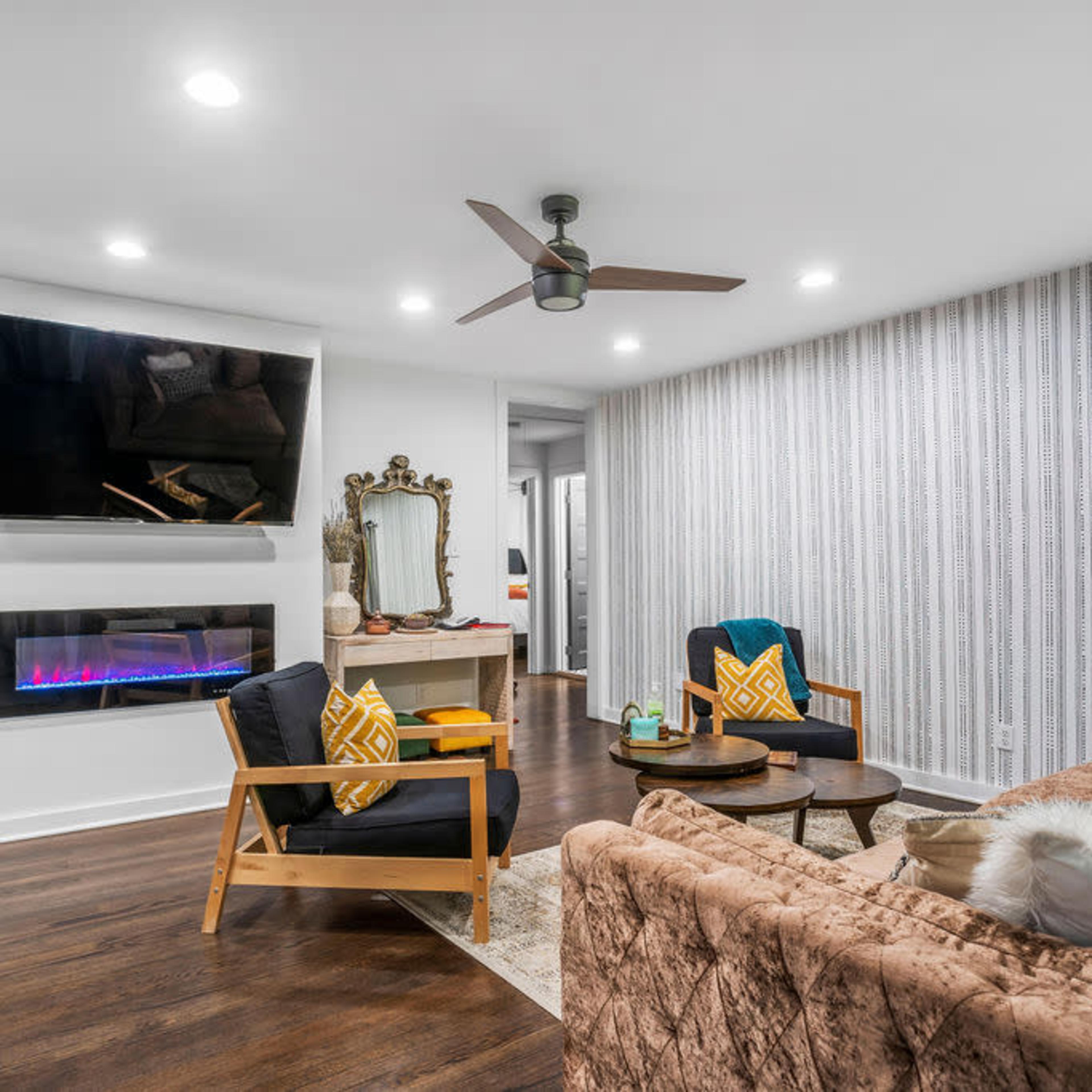 A modern living room features a wall-mounted TV, a sleek electric fireplace, and a mix of seating arrangements with decorative pillows.
