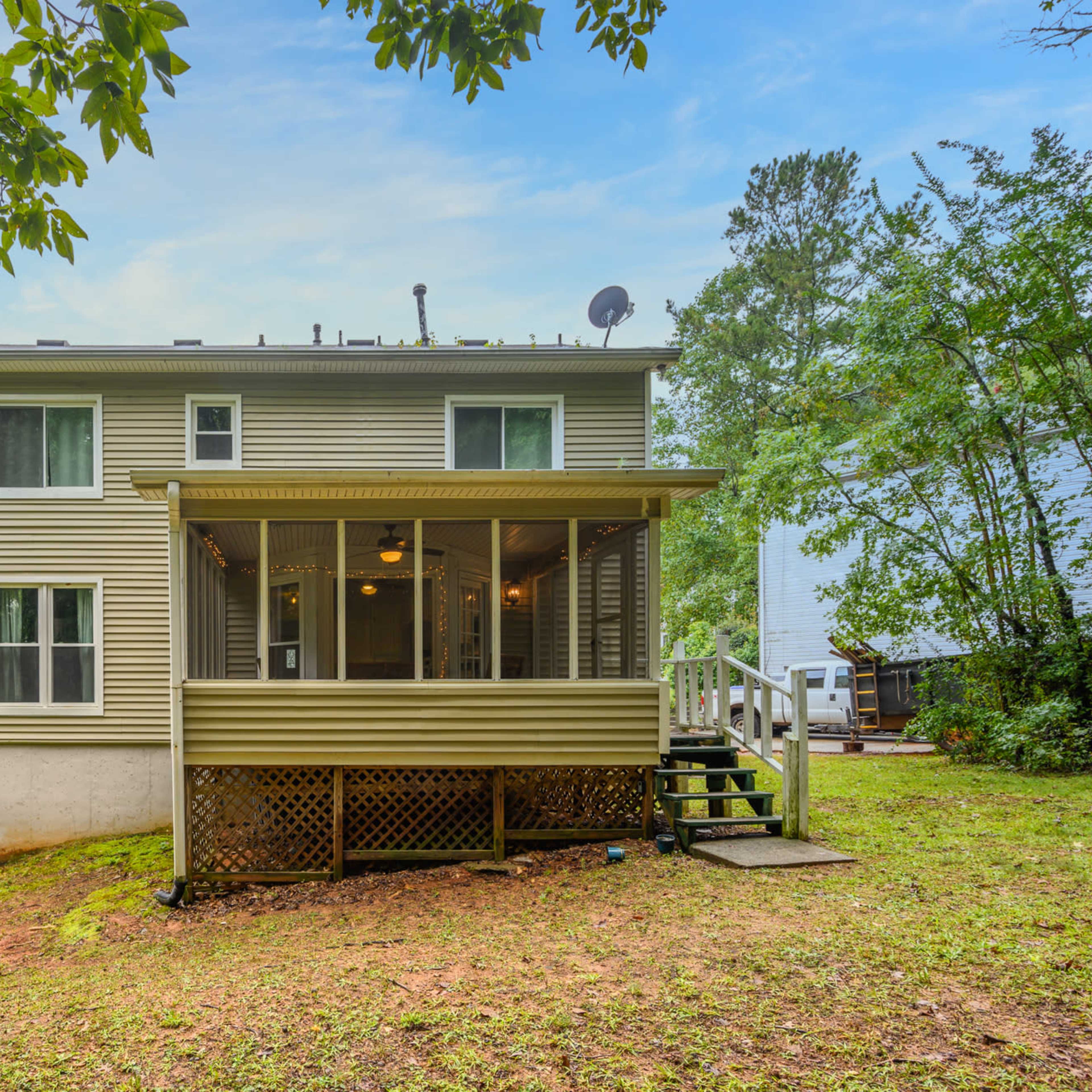 A two-story house with a screened porch sits on a grassy slope, surrounded by trees.