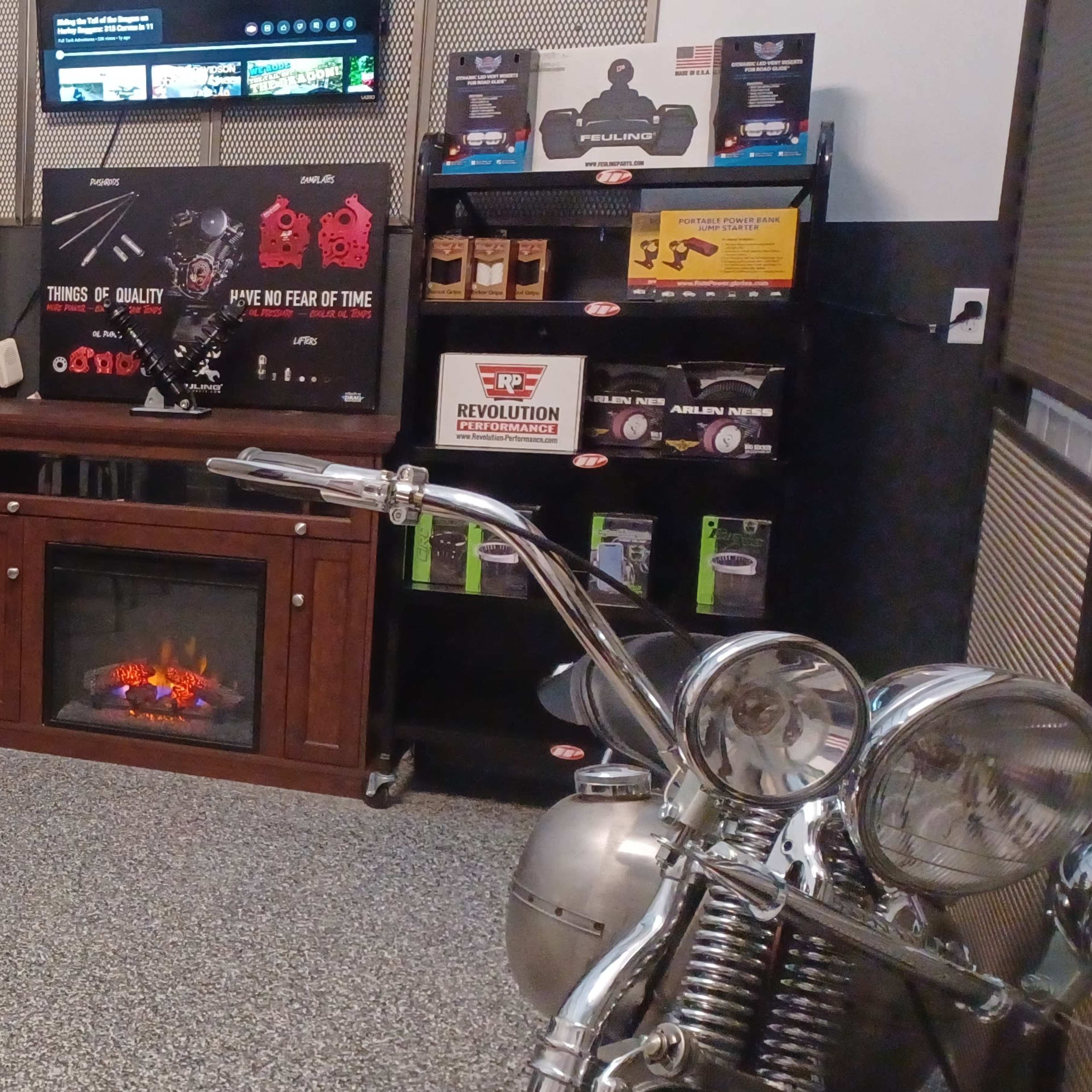 A garage setup featuring a wooden table, metal bar stools, a decorative electric fireplace, a display shelf with various products, and a motorcycle on the side.