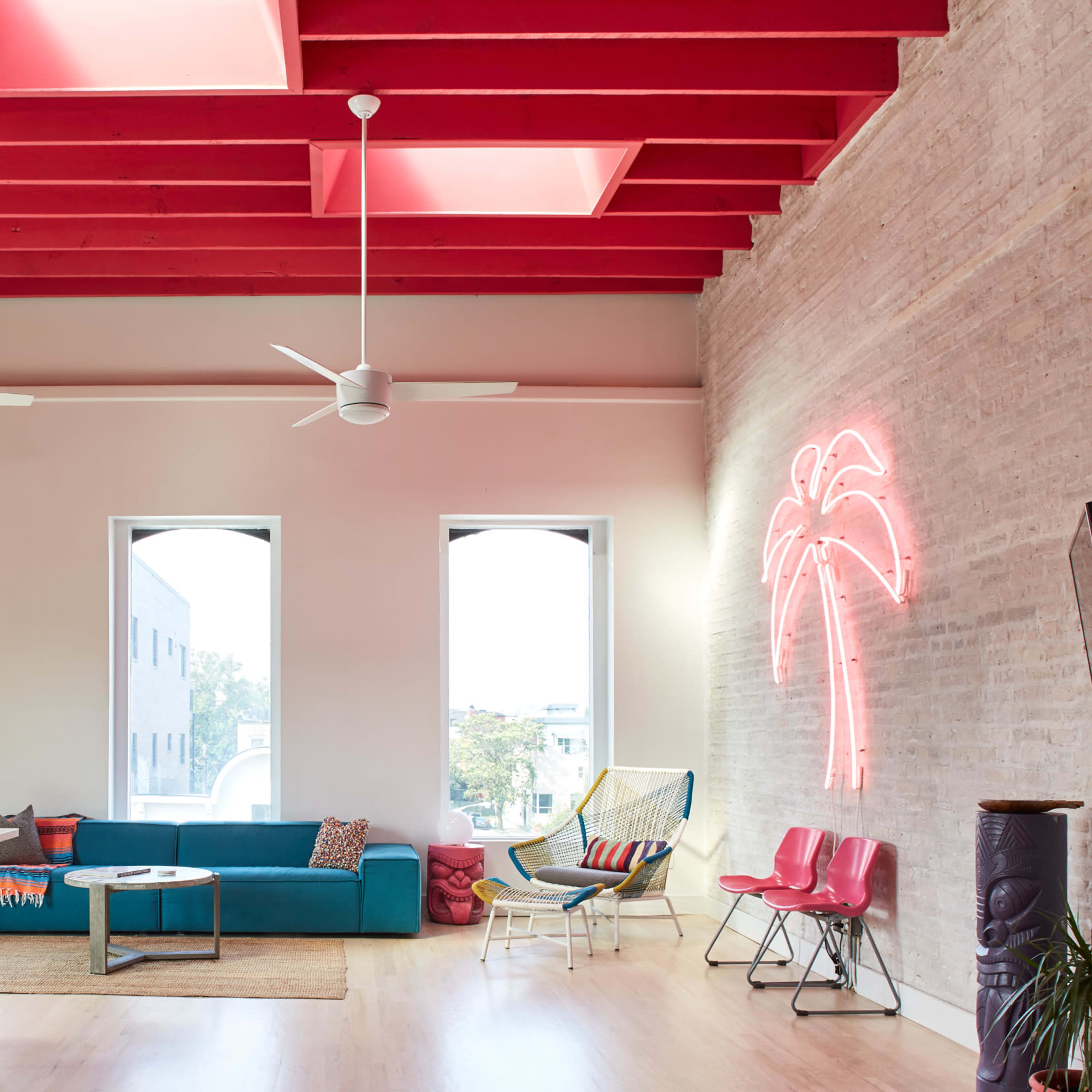 A modern, brightly lit living space with a turquoise sofa, a neon palm tree light, and colorful chairs under a ceiling with red beams.