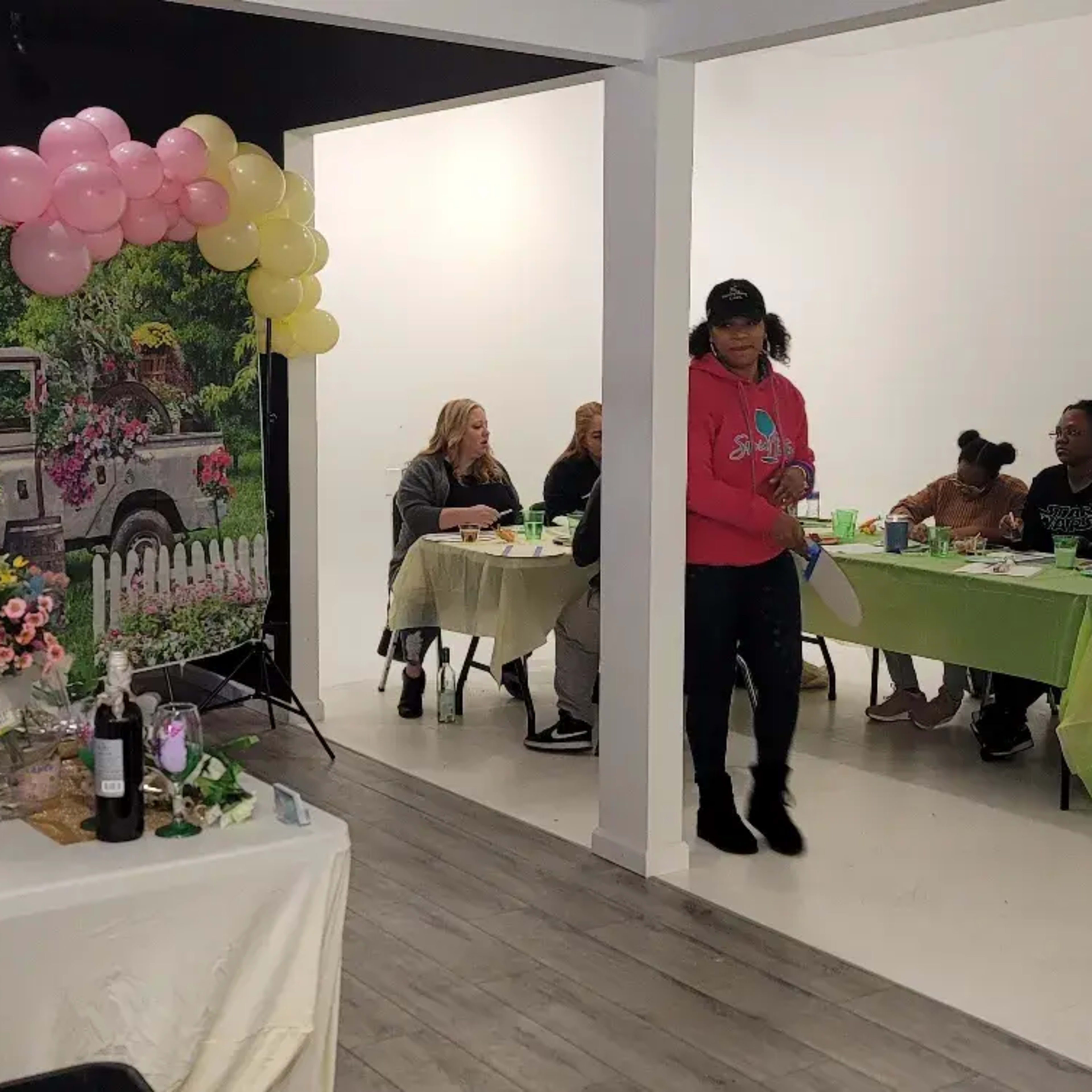 A group of people sits at tables in a room decorated with a floral backdrop and balloons, while one individual stands and interacts with others.