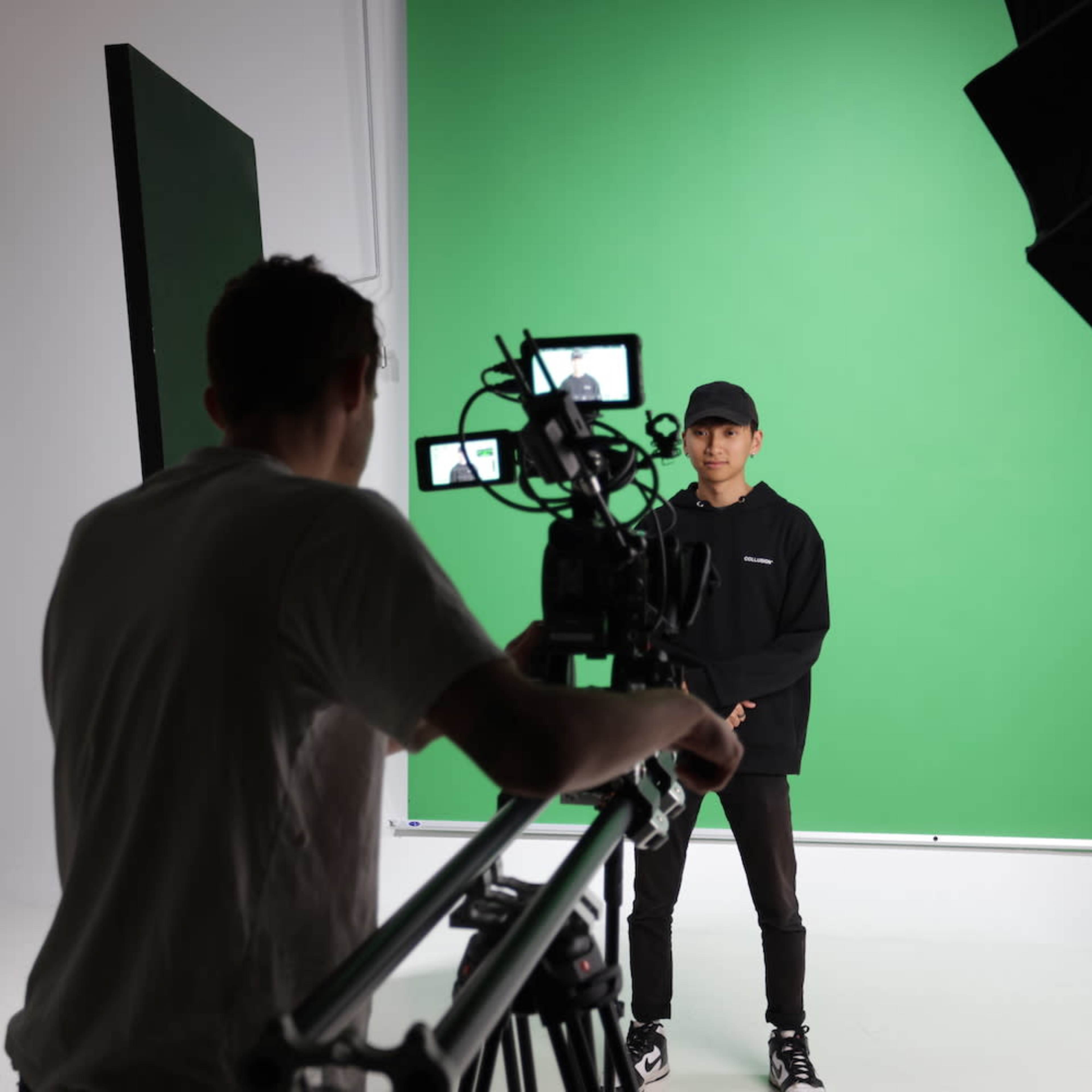 A person stands in front of a green screen while being filmed by a camera operator.