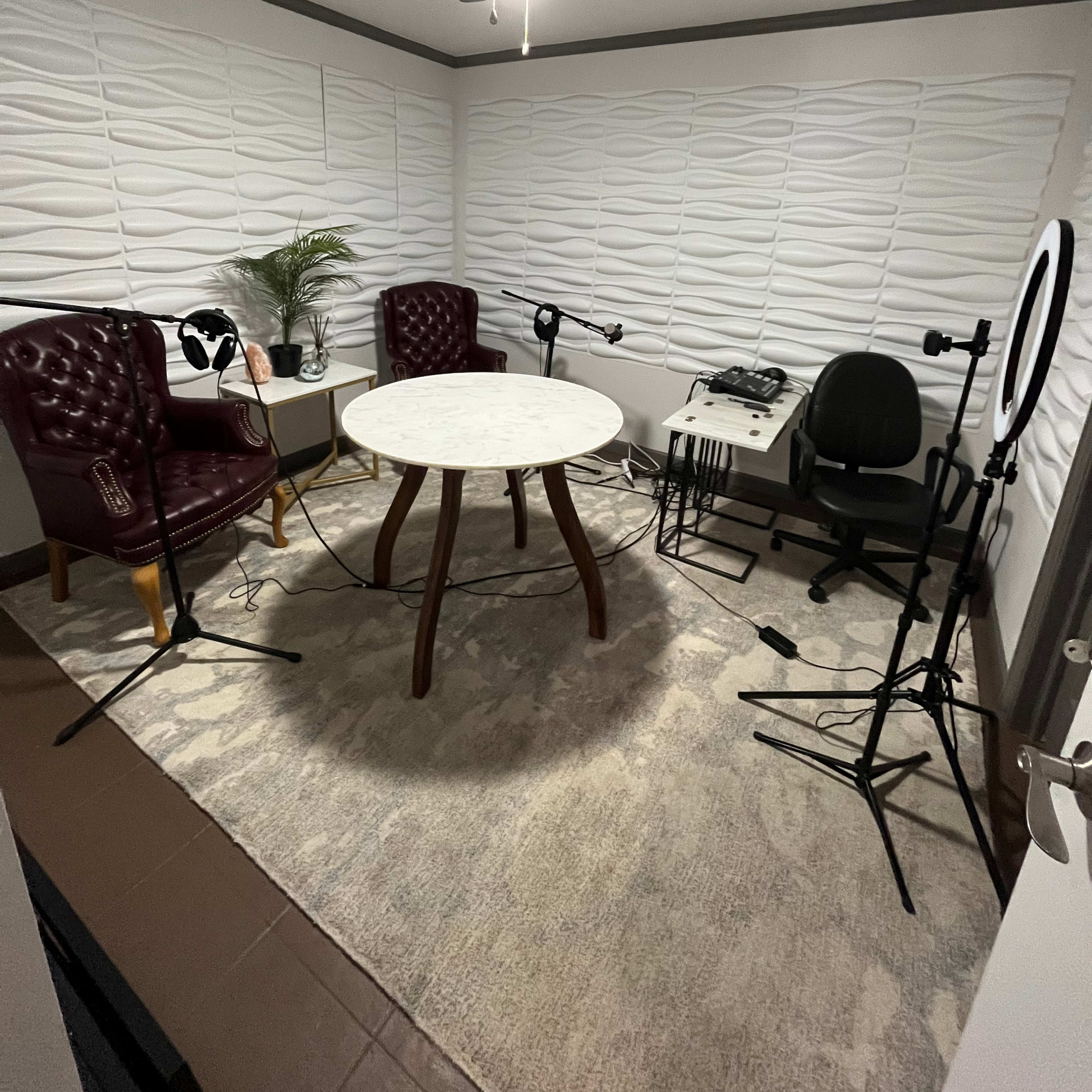 A small, well-furnished recording studio with two chairs, a round table, microphones on stands, and a ring light.