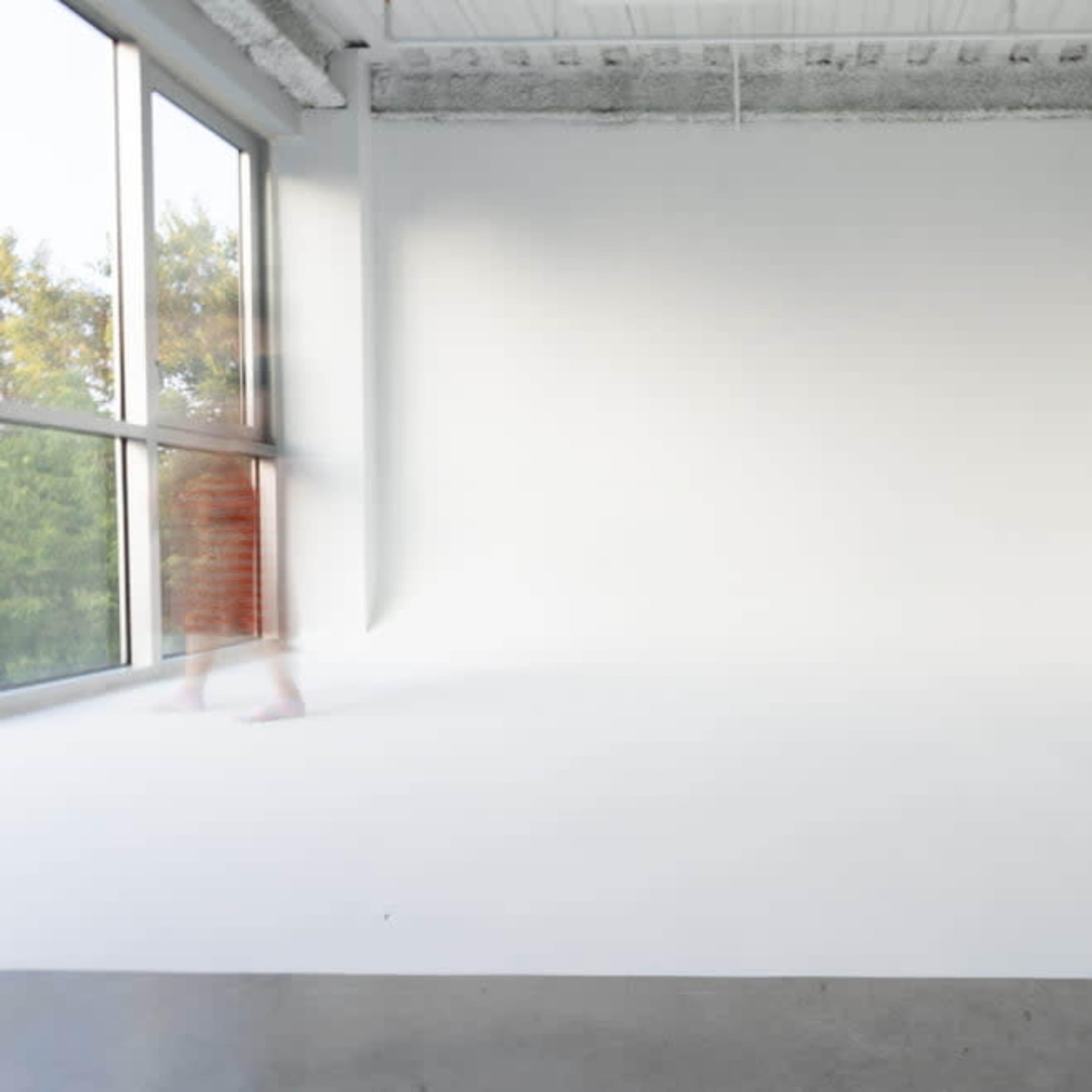 A person walks through an empty, well-lit room with white walls and a large window.