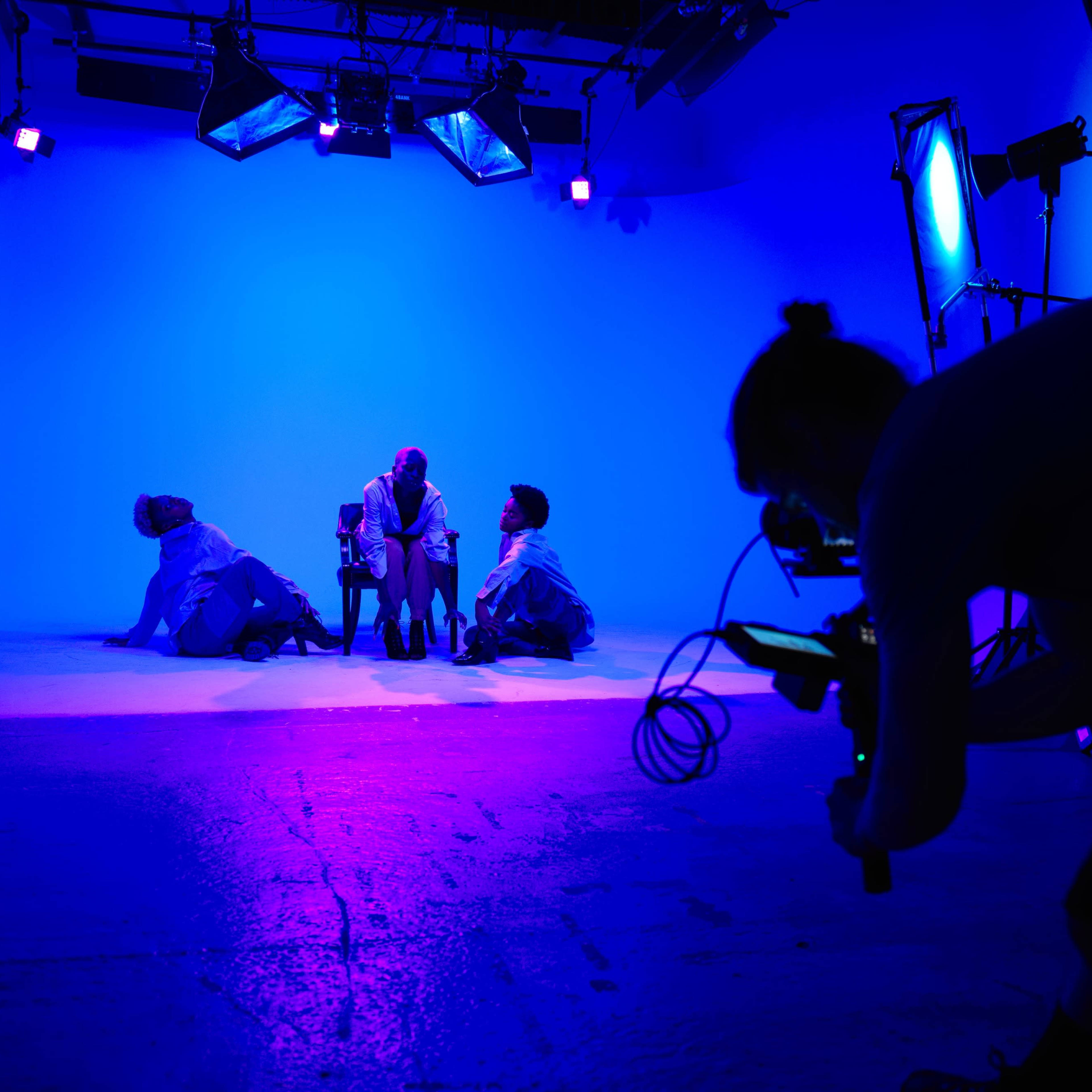 A person films a group of three performers in a brightly lit blue studio setting.