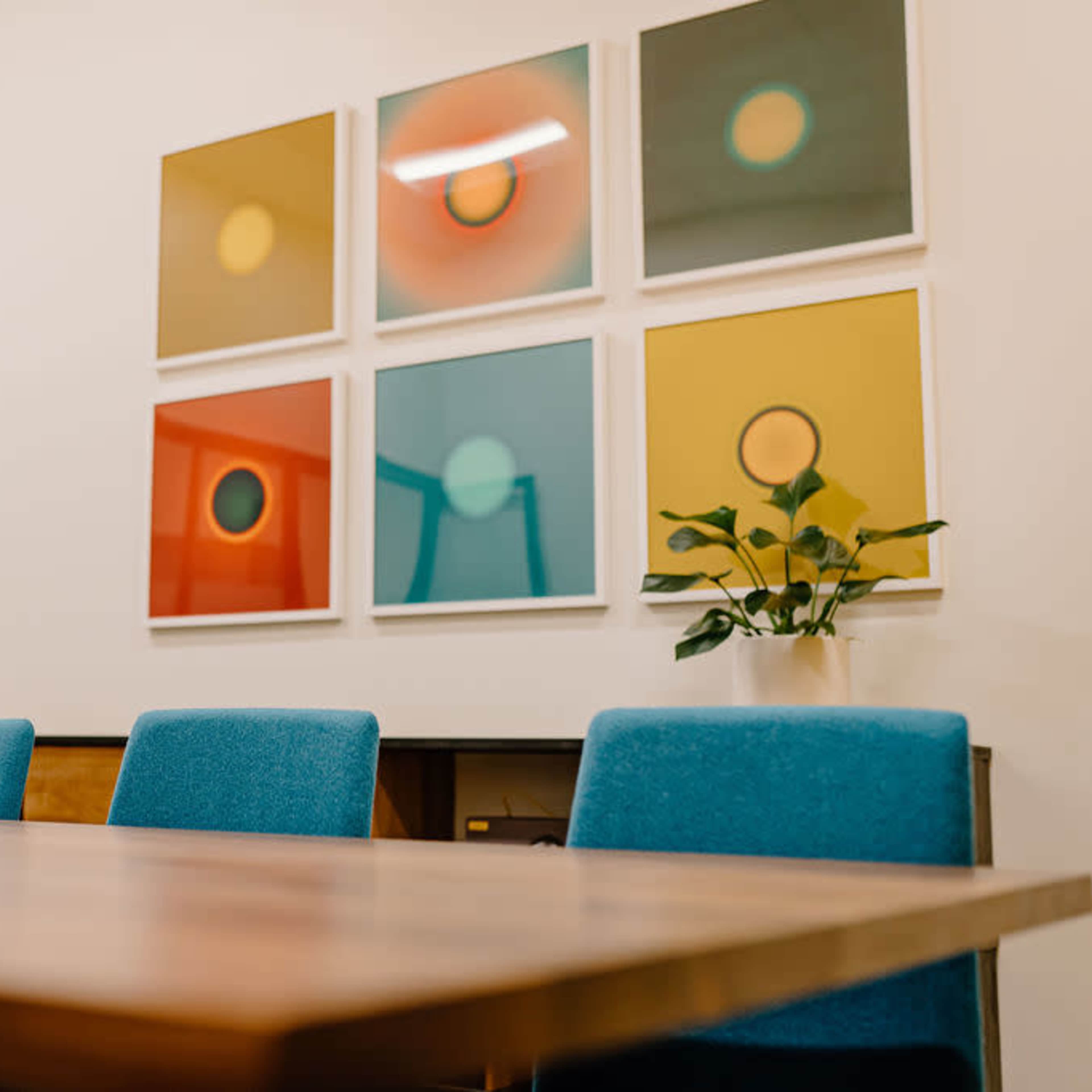 A conference room features a wooden table surrounded by blue chairs and colorful framed artwork on the wall.