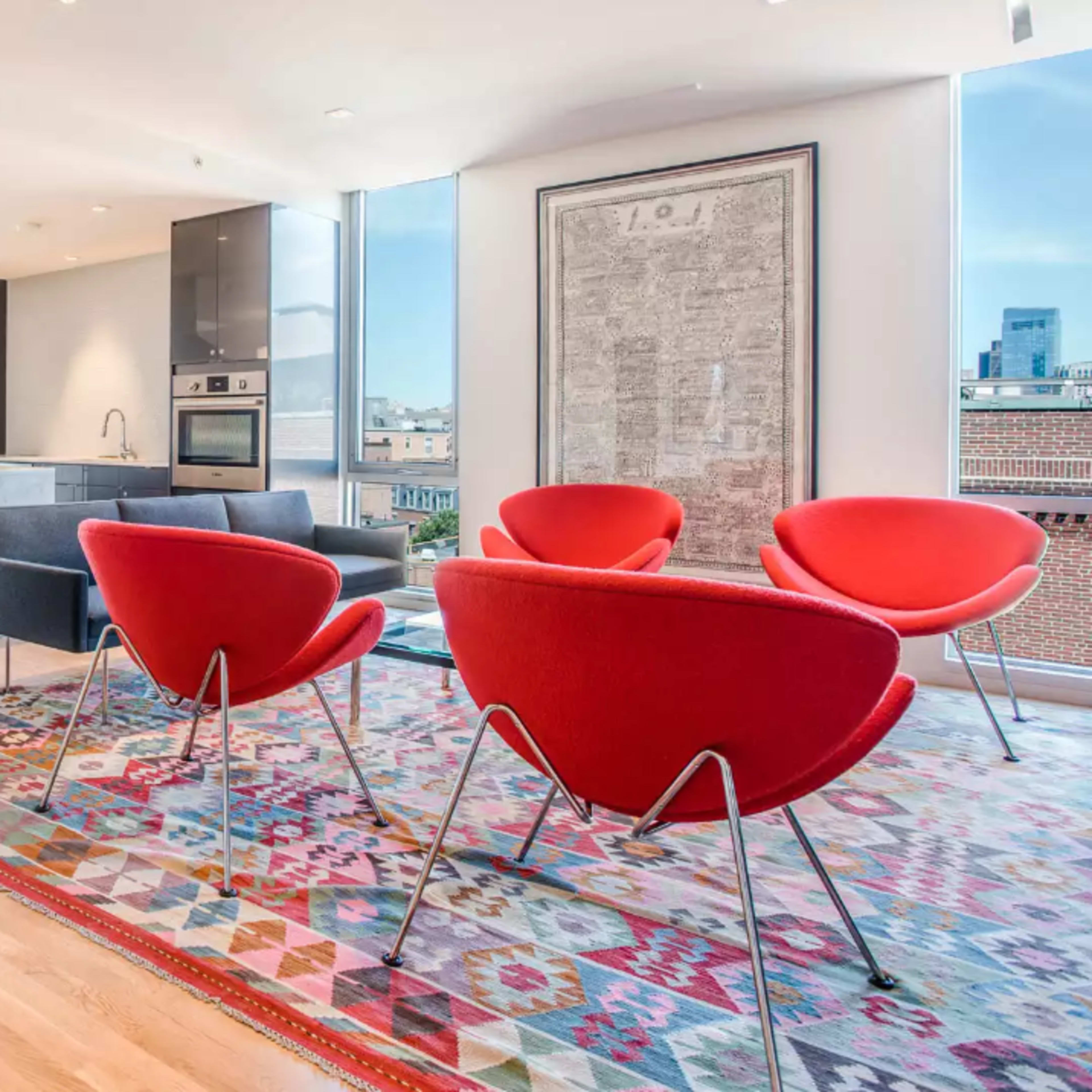 A modern open-concept living space featuring a kitchen, dining area, and seating area with bright red chairs on a patterned rug.