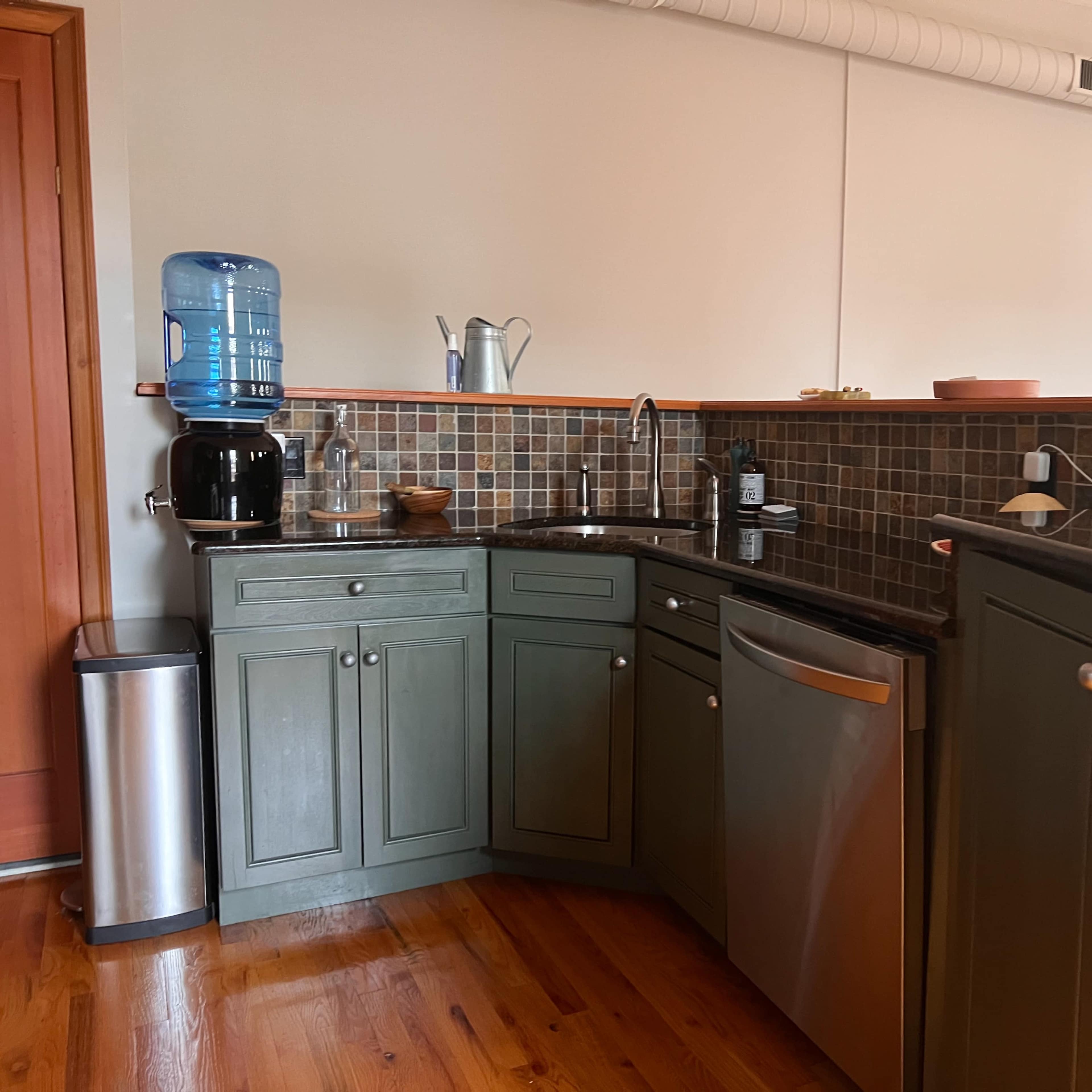 A kitchen with green cabinets, a stainless steel dishwasher, and a water dispenser next to a sink, all arranged along a tiled backsplash.