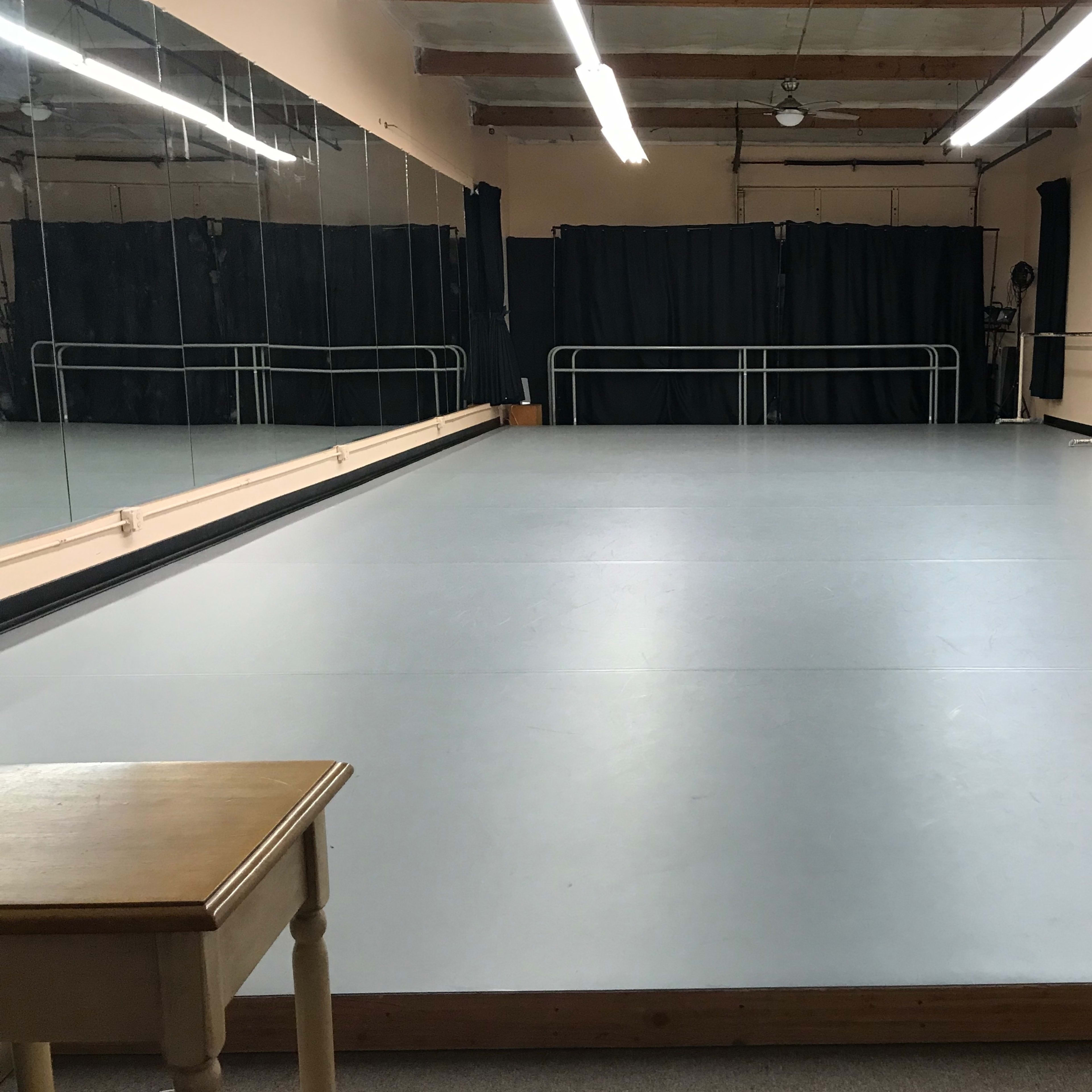 An empty dance studio with a gray floor, mirrored wall, and curtain-covered windows.