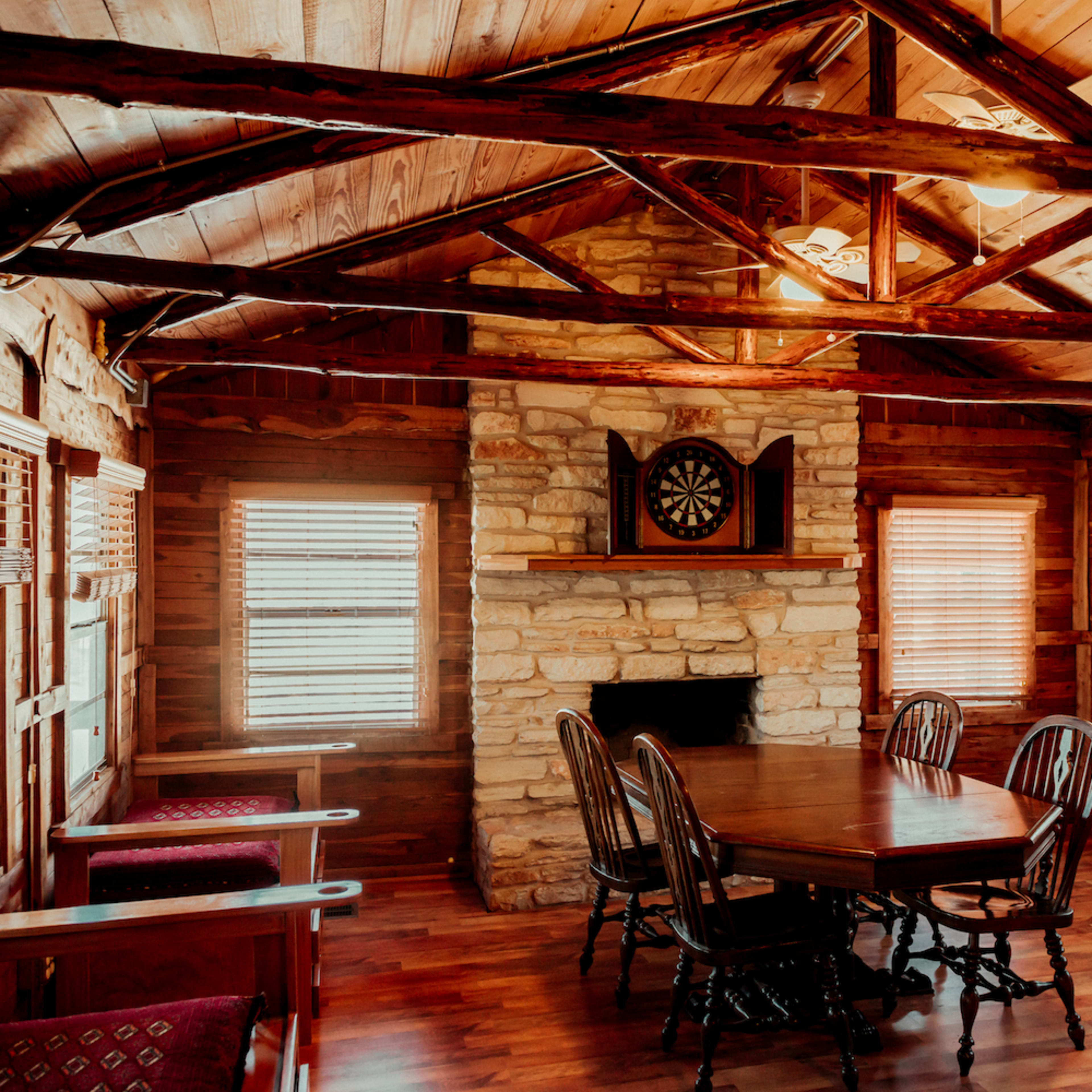 A rustic interior of a cabin with wooden walls and ceiling beams, featuring a stone fireplace, a round dining table, and several chairs.