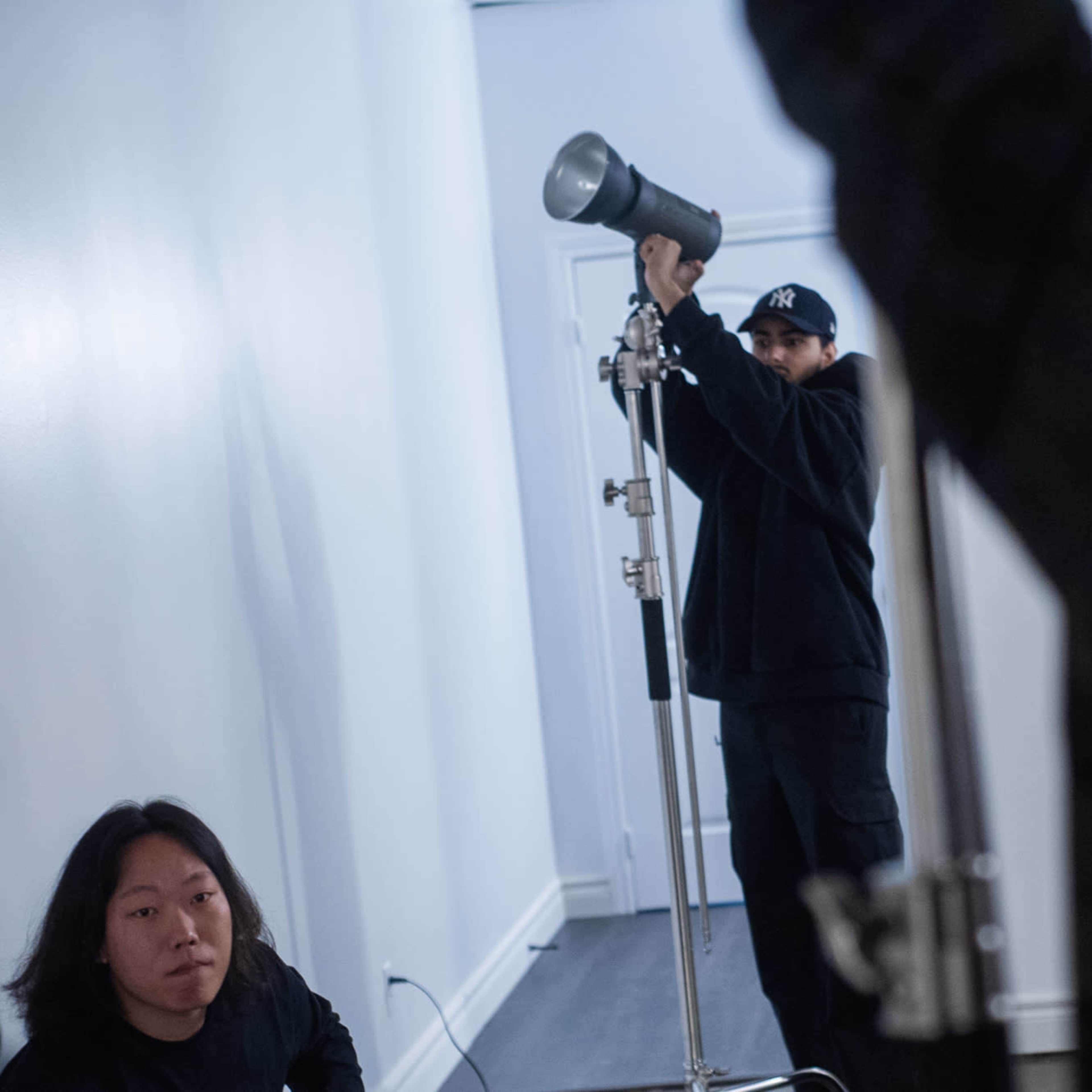 Two individuals are positioned in a minimalistic studio setting, with one preparing a lighting setup and the other seated on the floor.