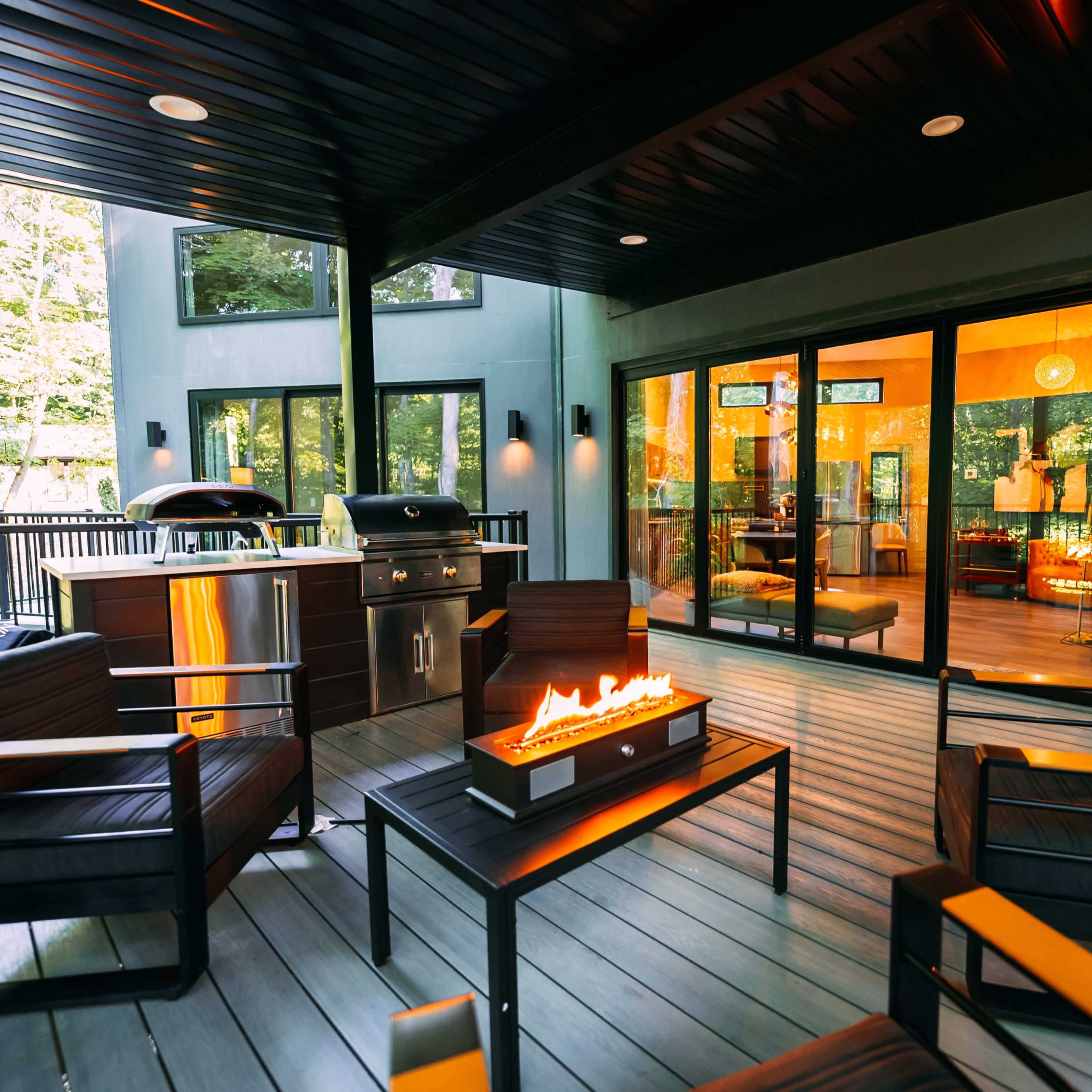 A modern outdoor seating area with a fire pit, grill, and large windows overlooking a cozy indoor space.
