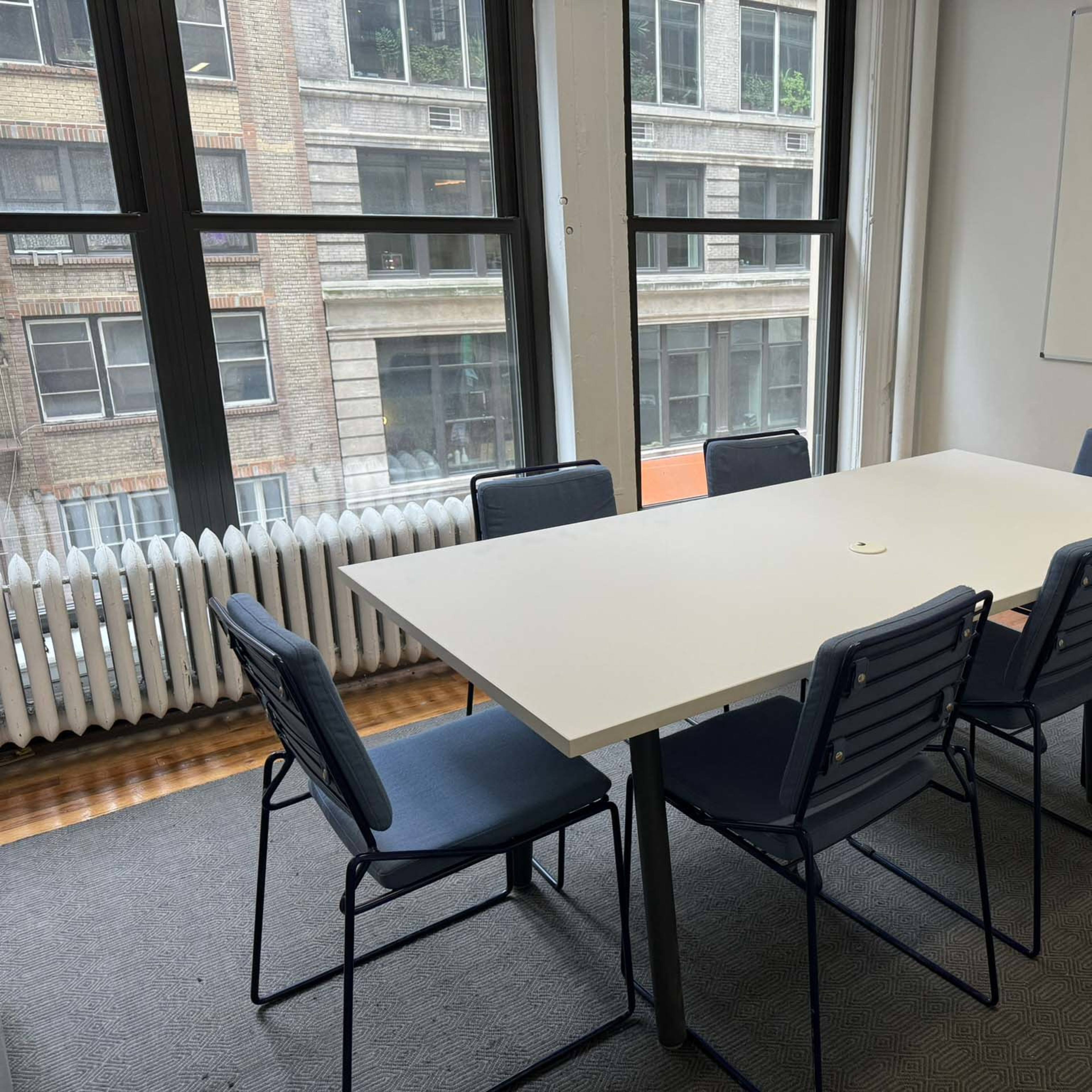 A conference room features a large white table surrounded by six chairs, with windows overlooking a city street.