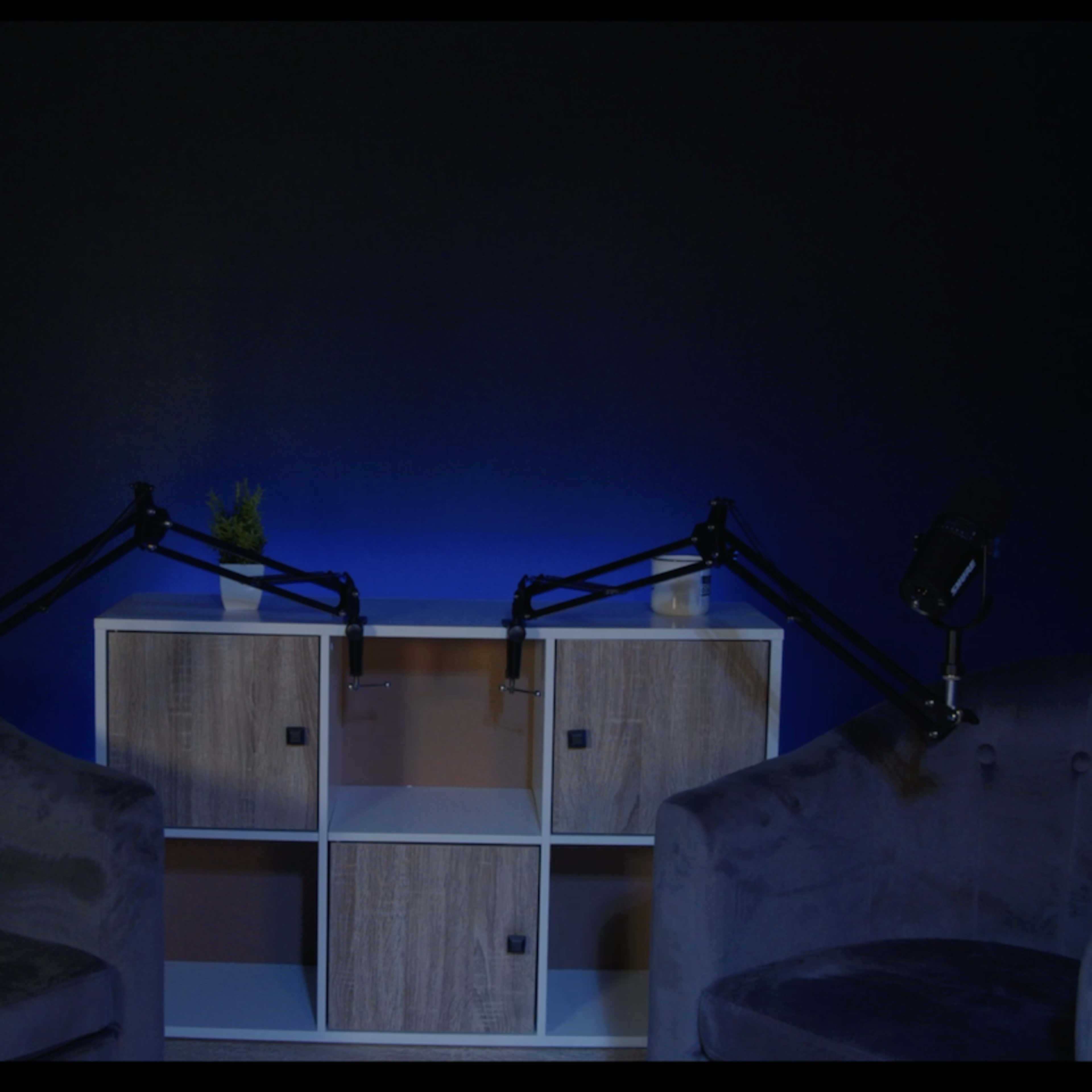 Two dark, plush armchairs are positioned facing each other in front of a shelf with blue lighting.
