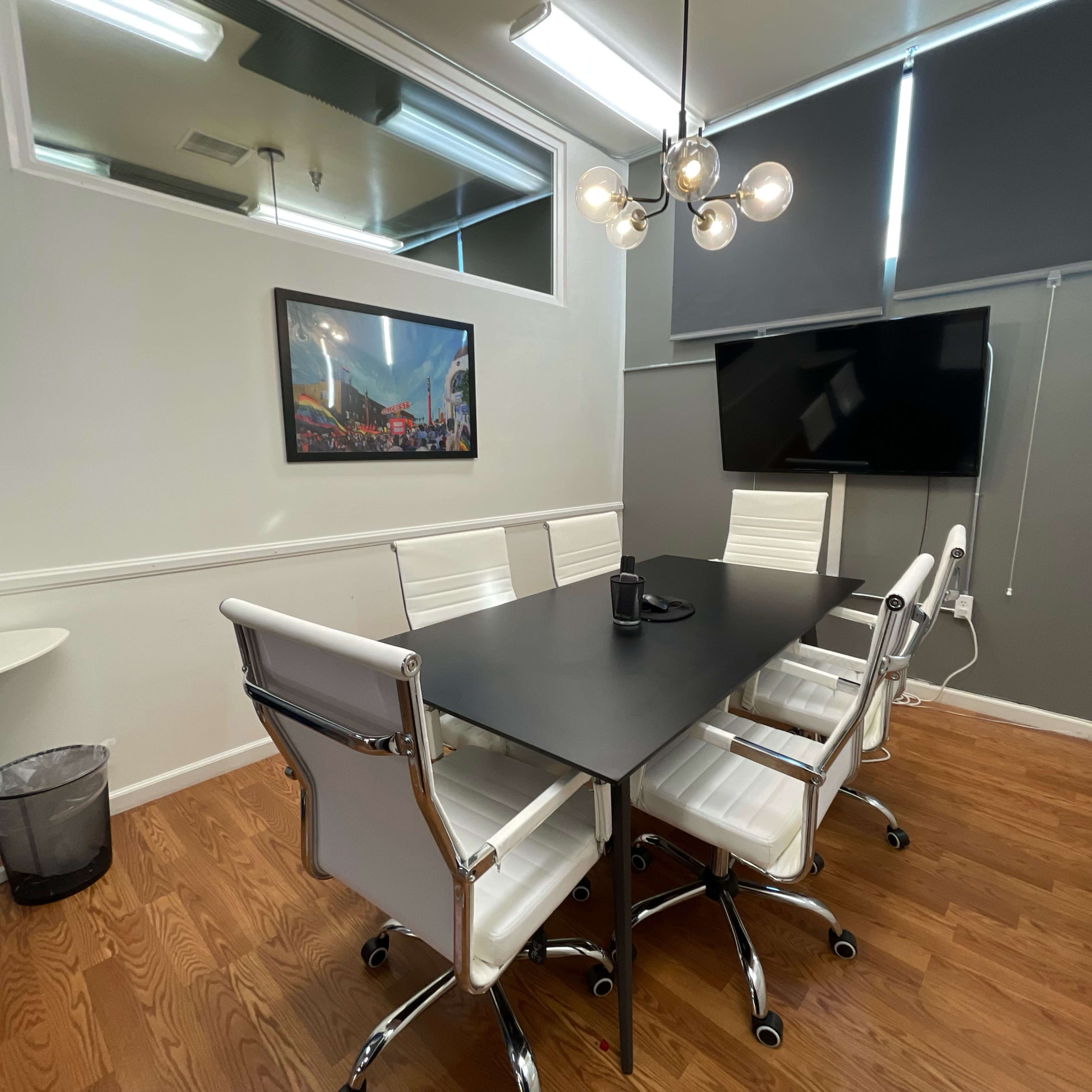 A conference room features a black table surrounded by six white chairs, with a wall-mounted screen and a framed picture.