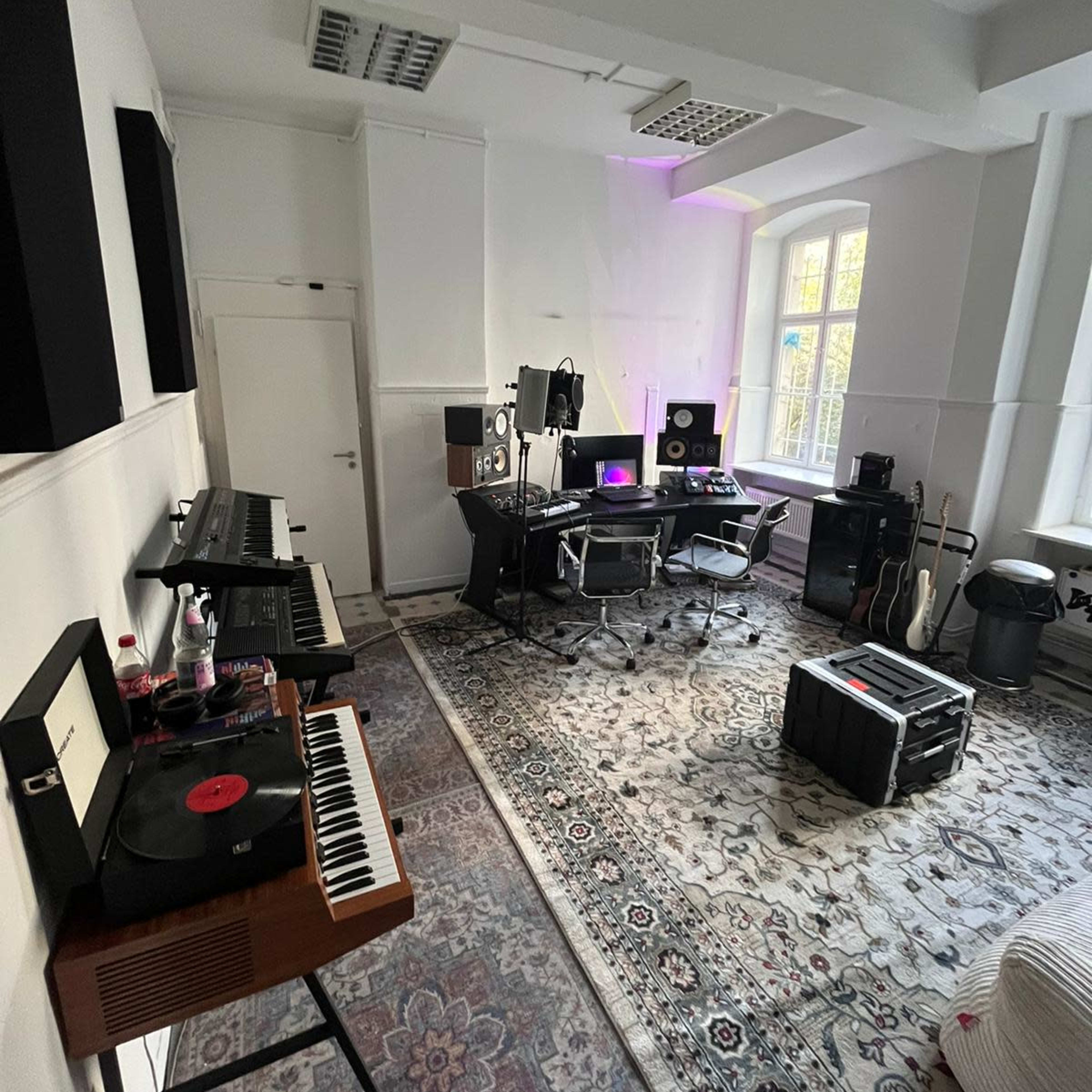 A music production studio featuring keyboards, recording equipment, a desk with monitors, and a large area rug.