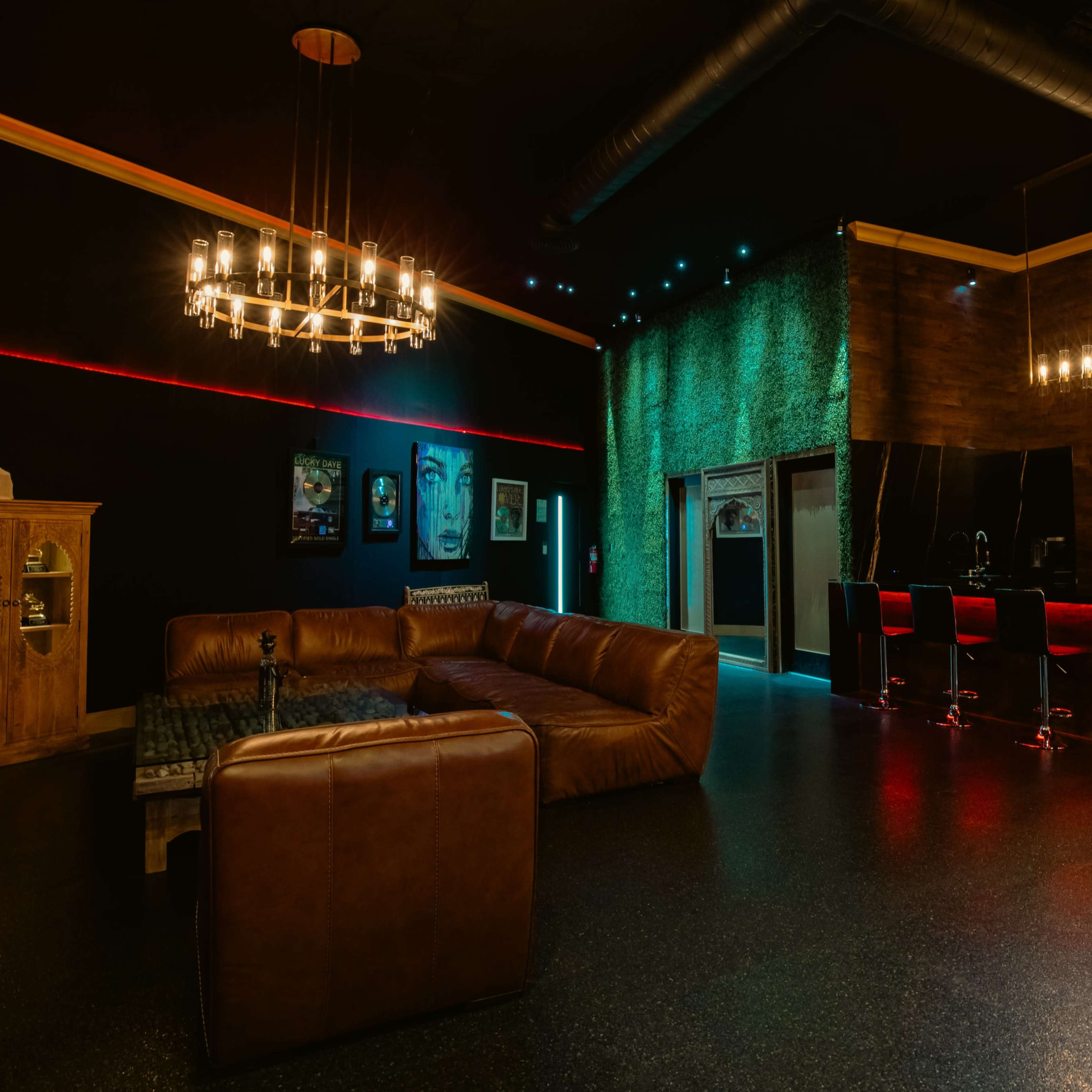 A modern interior space featuring a sectional leather sofa, a wooden cabinet, a bar area with high stools, and wall decor, illuminated by various light fixtures.