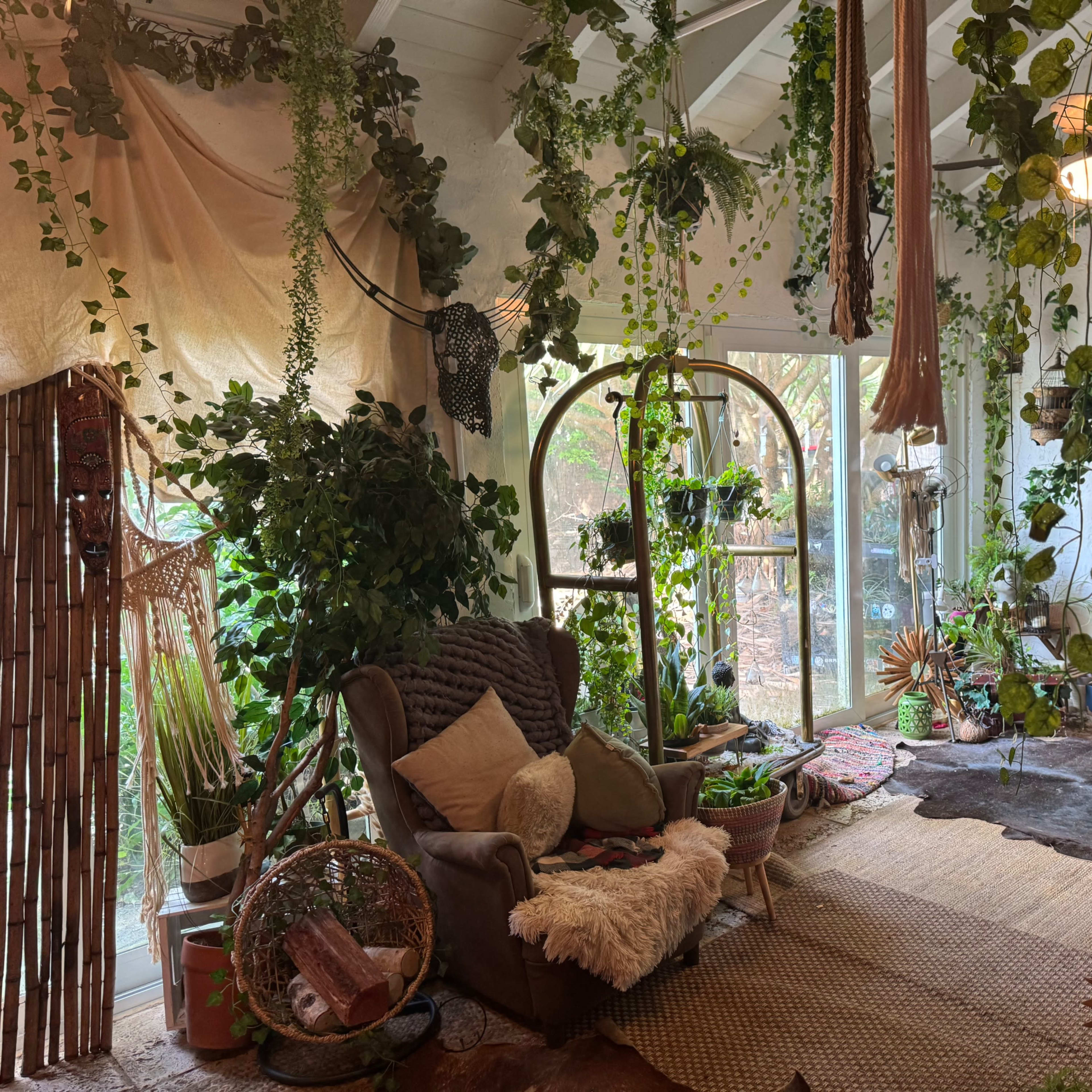 A cozy living space featuring a brown armchair surrounded by various plants, decorative items, and light filtering through large windows.