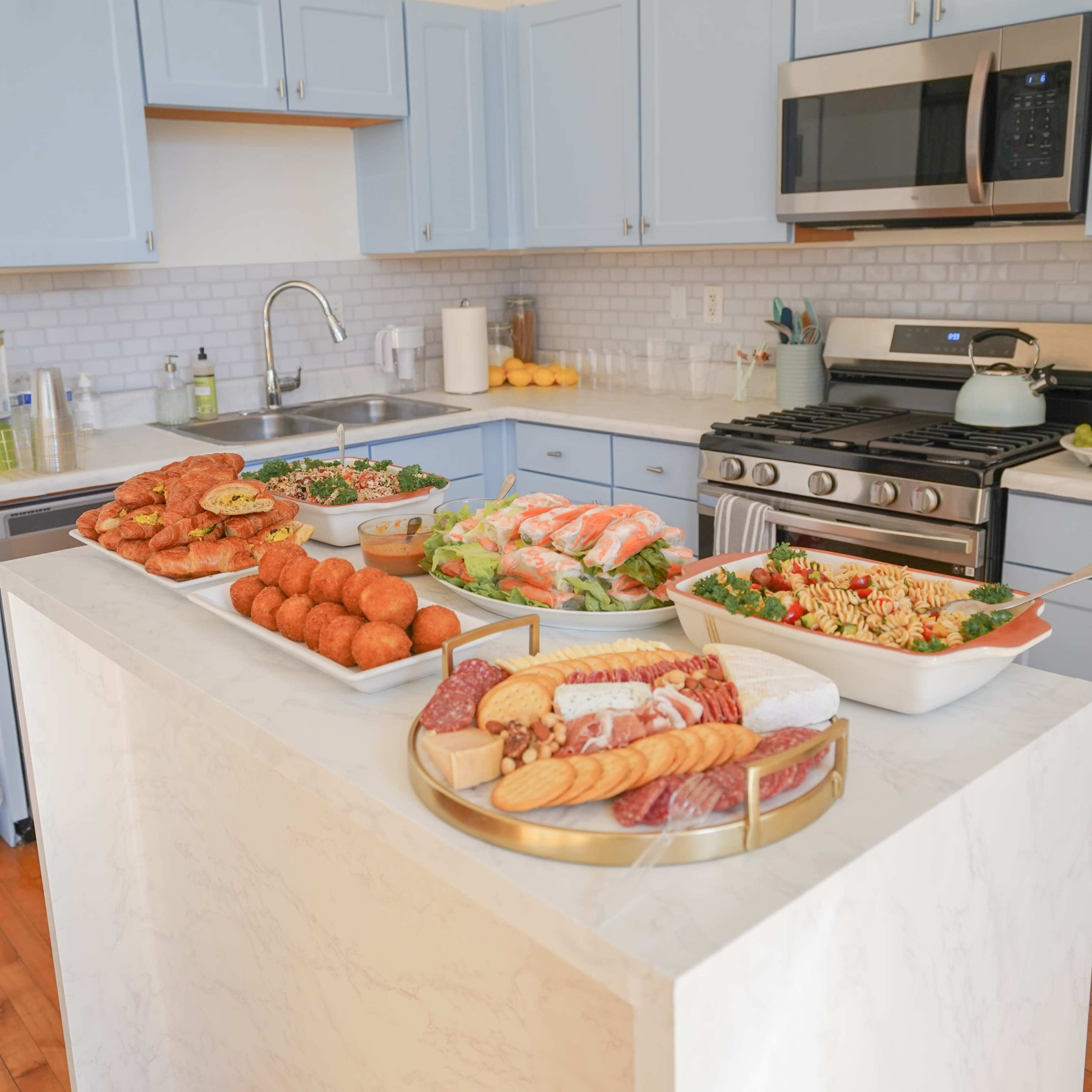 A kitchen island is filled with a variety of prepared foods and dishes, including salads, meats, and pastries, in a brightly lit kitchen with blue cabinetry.