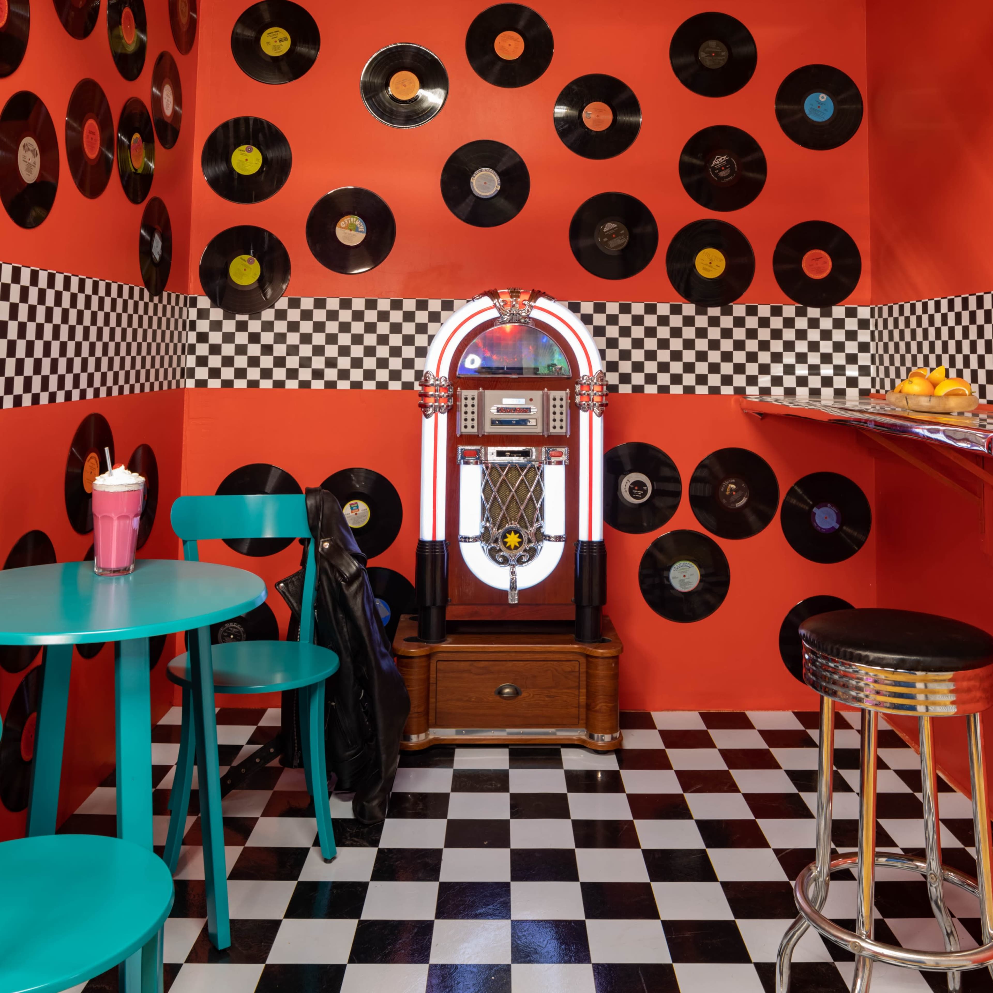 A small retro-themed room features a jukebox against a brightly colored red wall adorned with black vinyl records, with checkered flooring and a table set for two.
