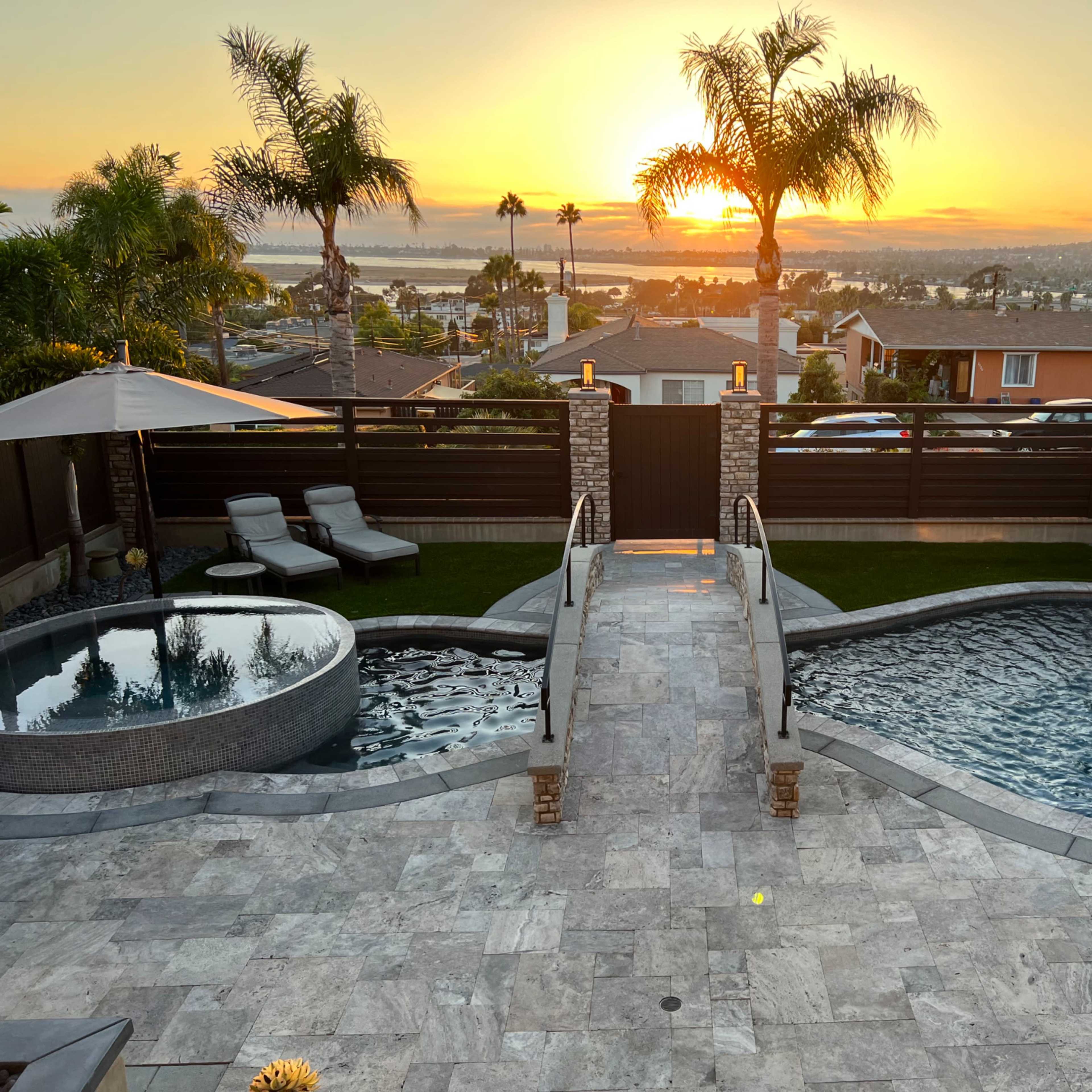 A backyard with a spa, swimming pool, and a sunset backdrop, framed by palm trees and a landscaped garden.