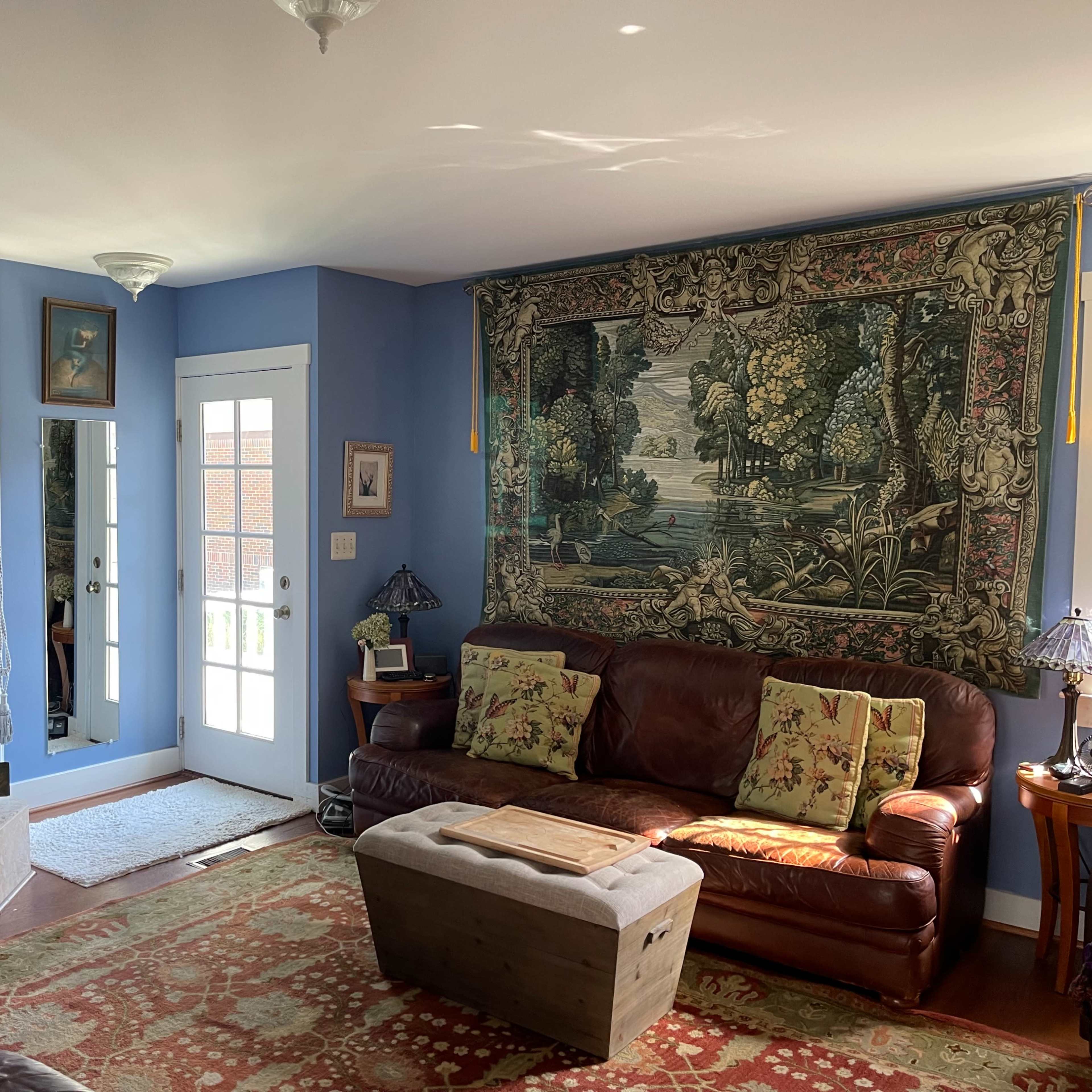 A living room with blue walls, a stone fireplace, a large tapestry on one side, and leather couches arranged around a wooden coffee table.
