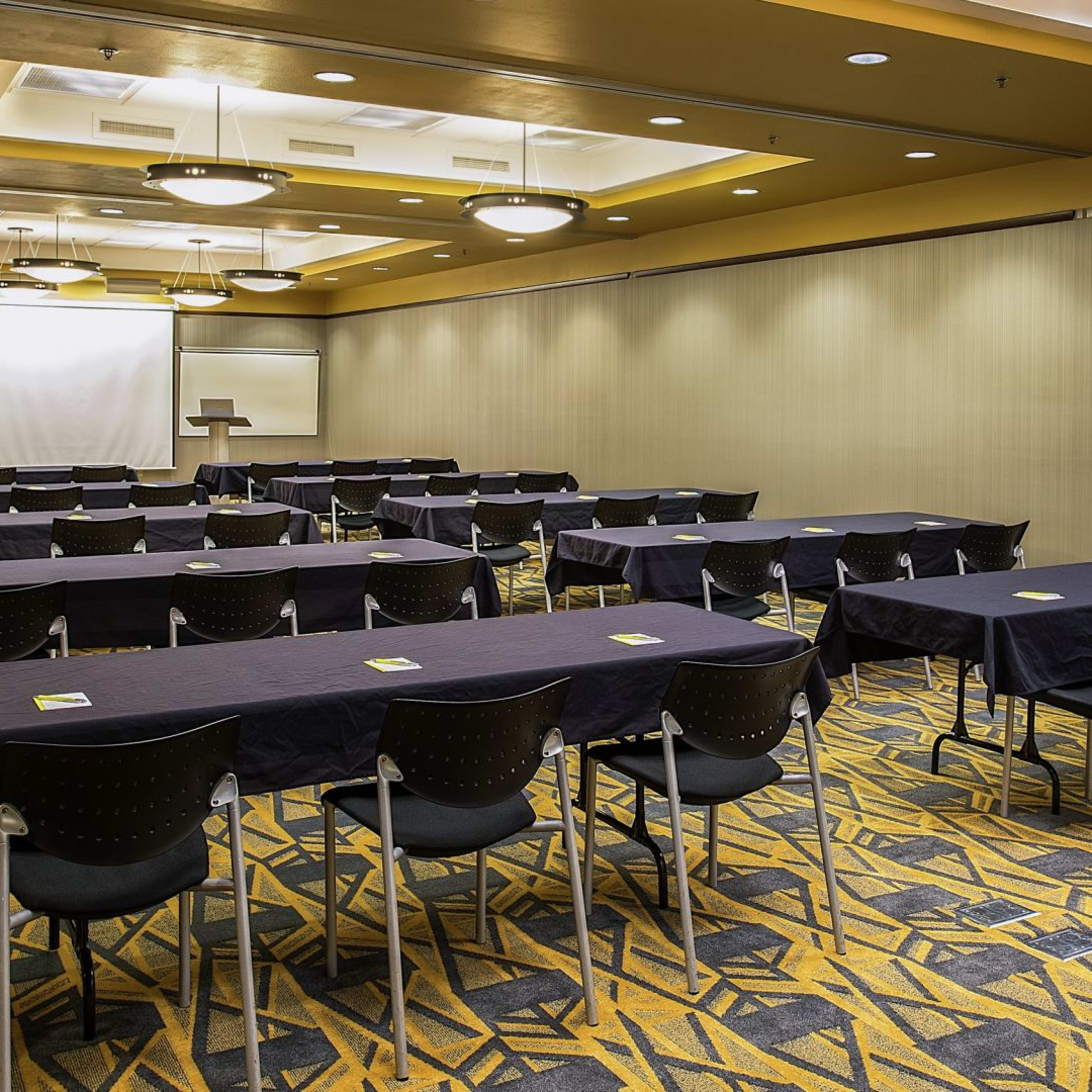 A conference room with multiple tables arranged in rows, each covered with black tablecloths and facing a projection screen.
