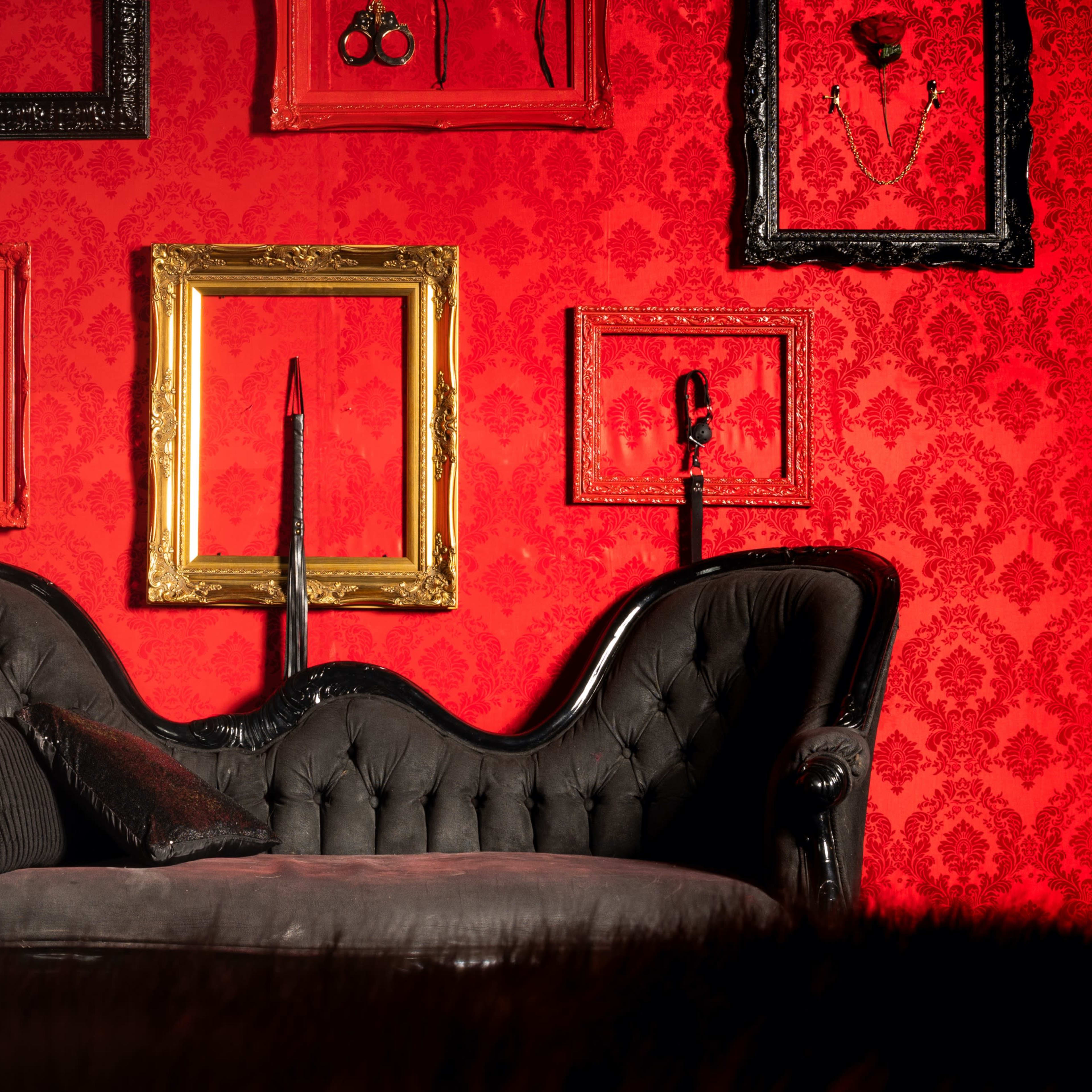 A black velvet couch positioned against a red damask wall adorned with ornate picture frames and decorative sculptures.