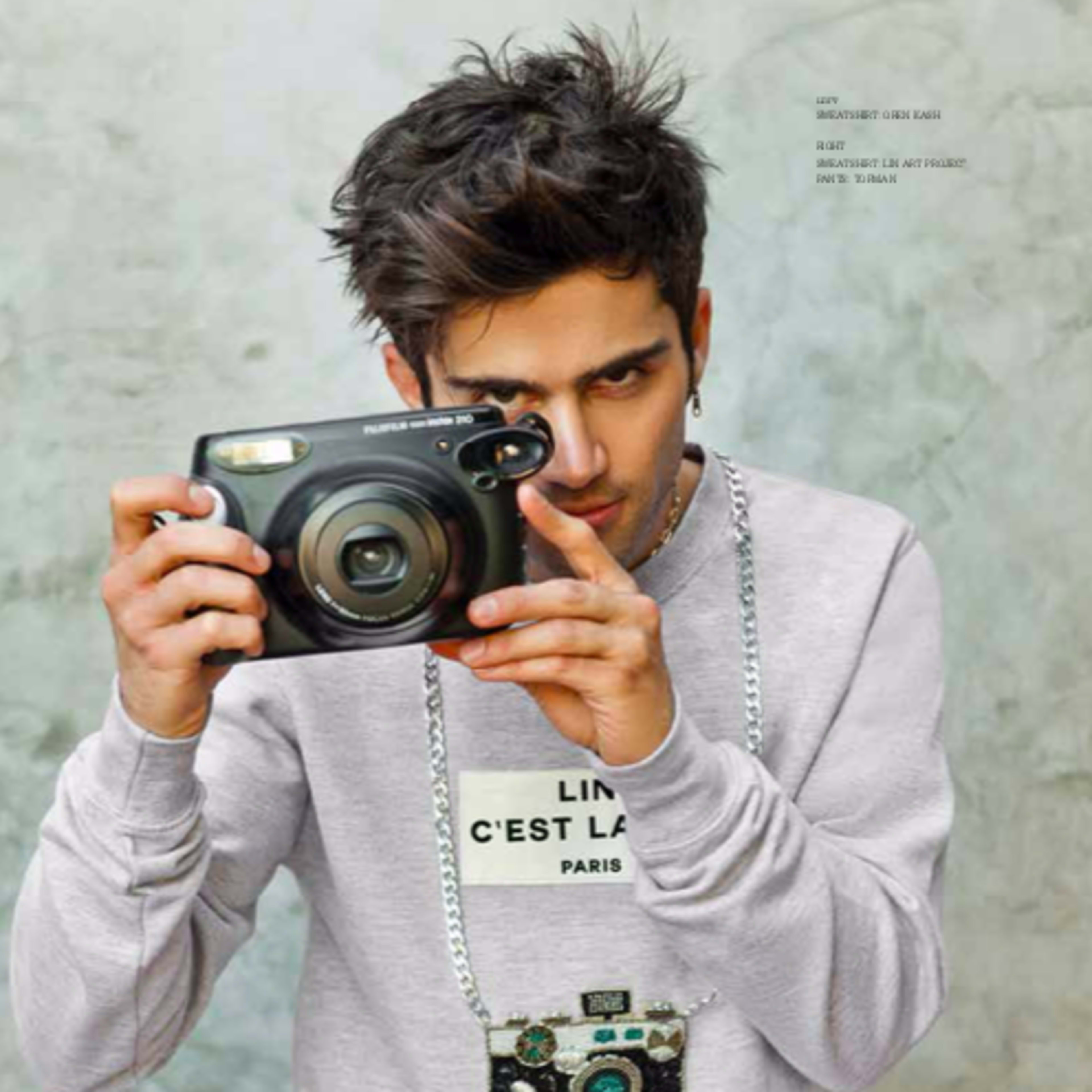 A young man wearing a gray sweatshirt with a camera graphic is holding a camera and posing for the photo.