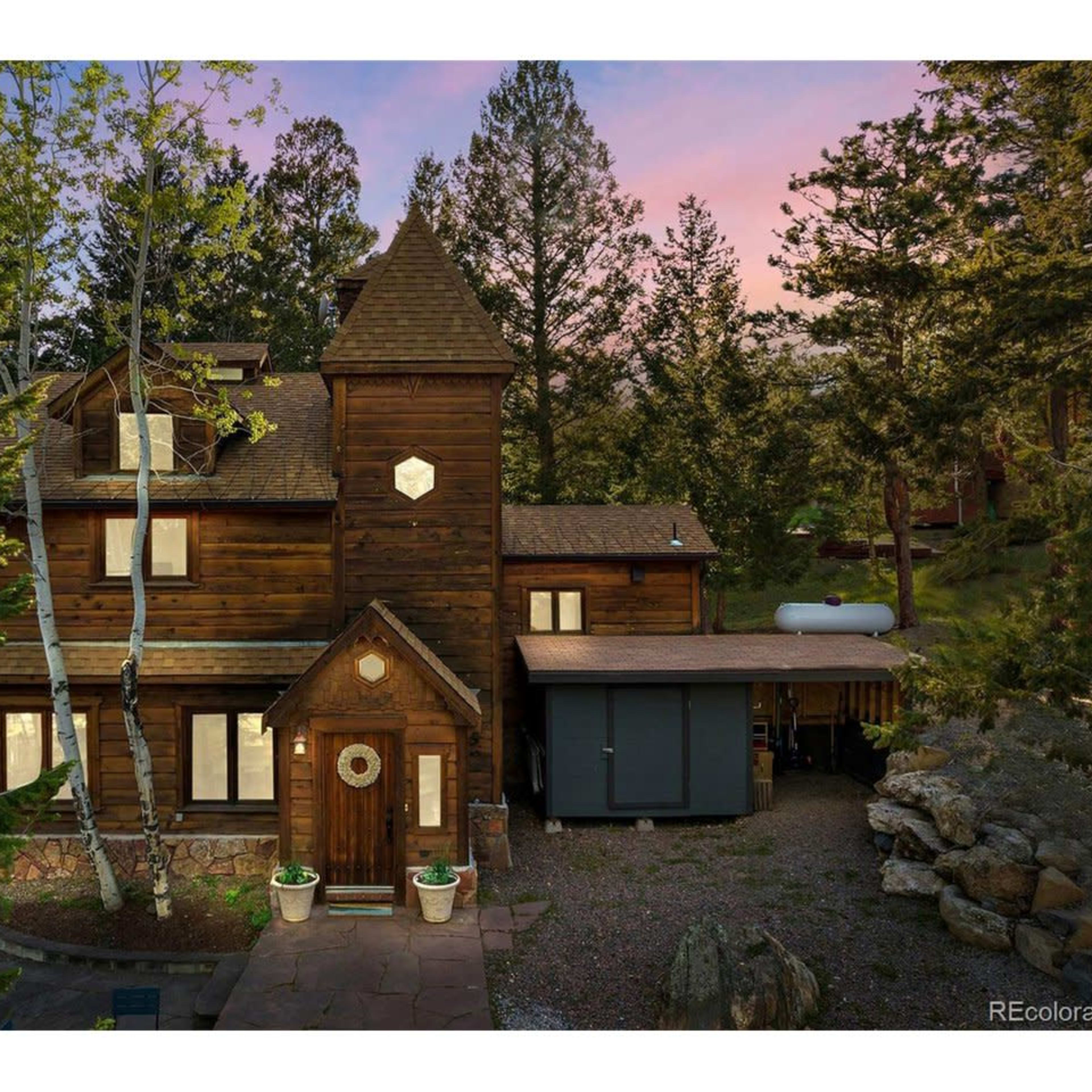 A multi-story wooden house surrounded by trees, featuring a unique tower and a stone pathway leading to the entrance.