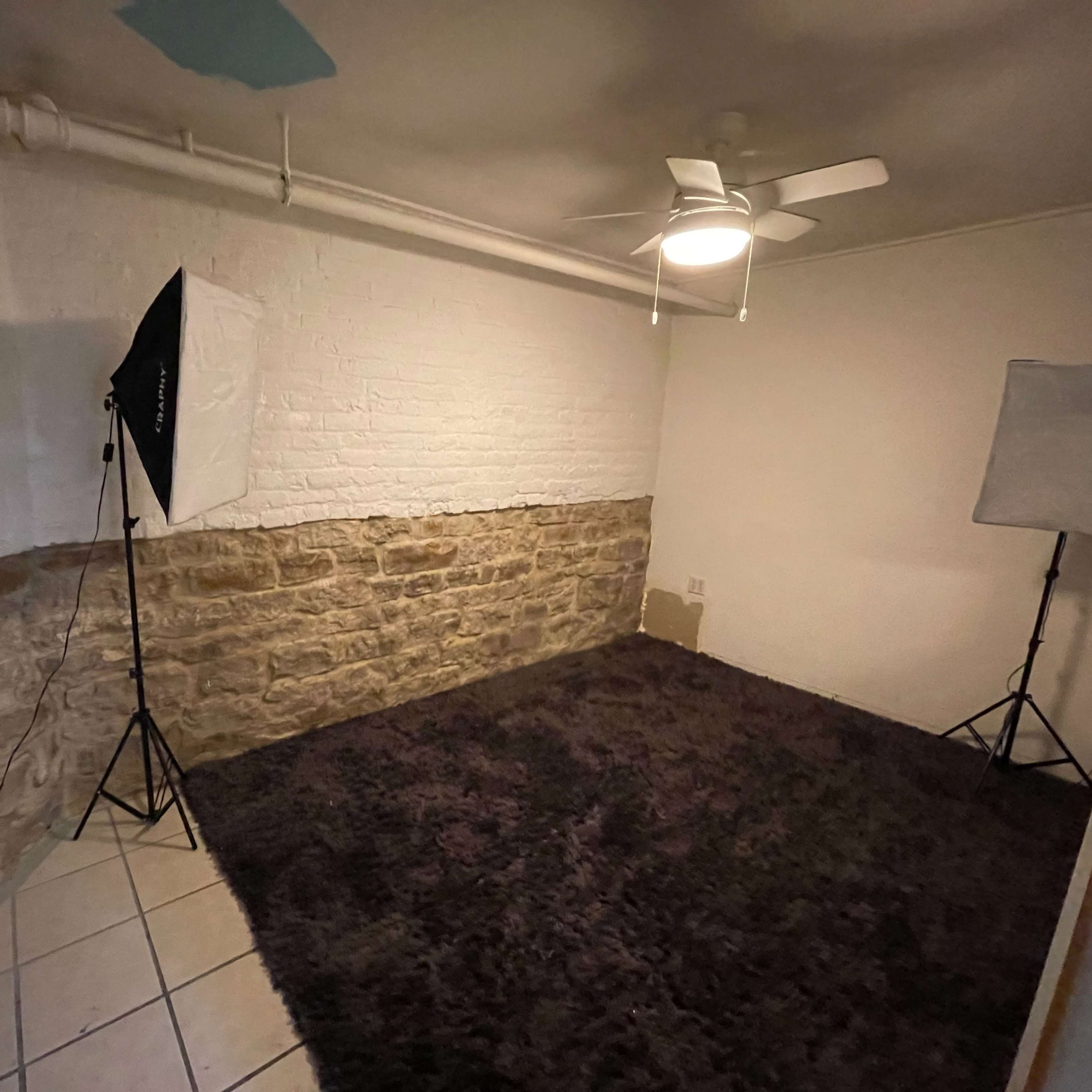 A sparsely furnished room with a stone wall on one side, a ceiling fan, and photography lights set up in the corners.
