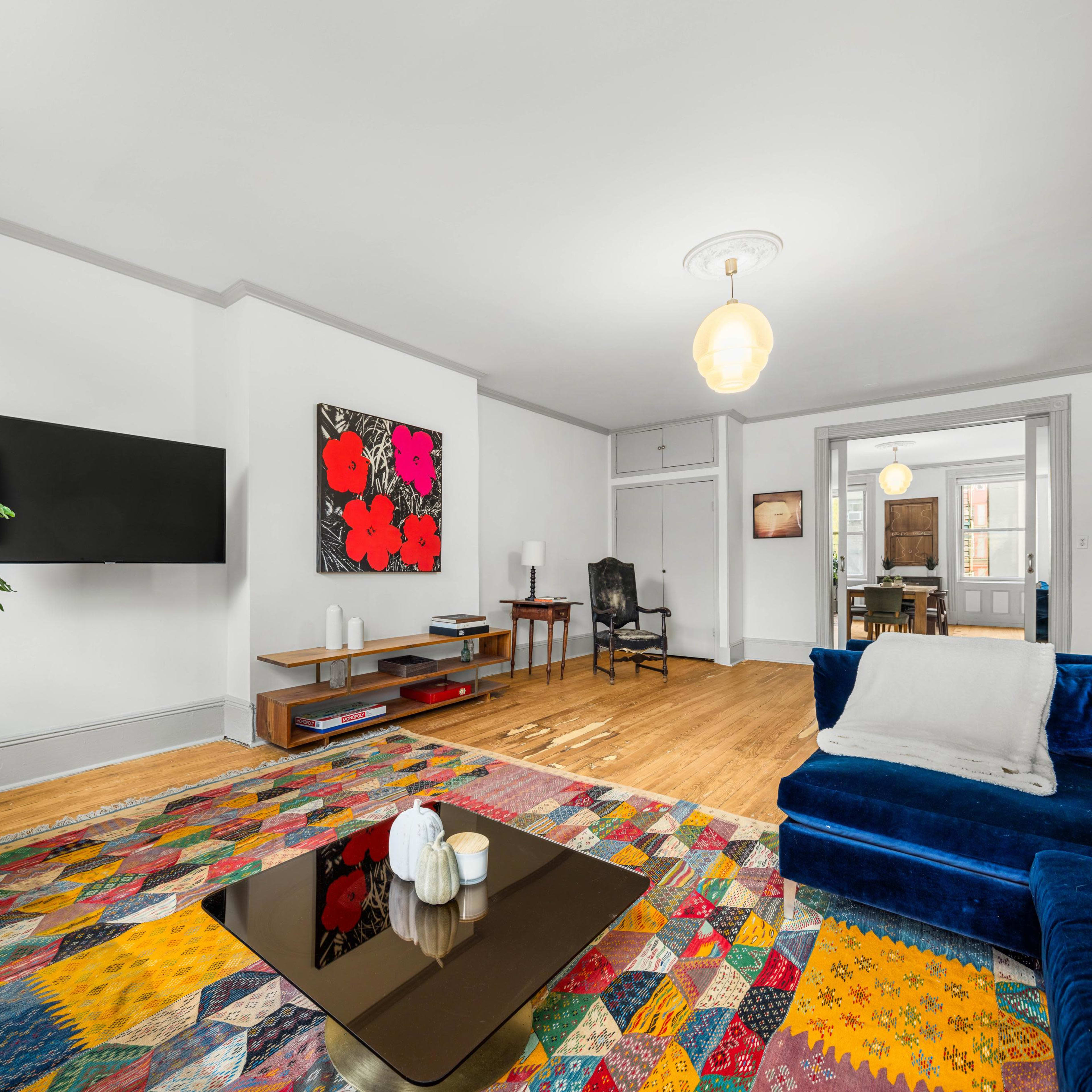 A living room features a colorful patterned rug, a blue sofa, a coffee table, and a wall-mounted television, with an artwork and a potted plant in the background.
