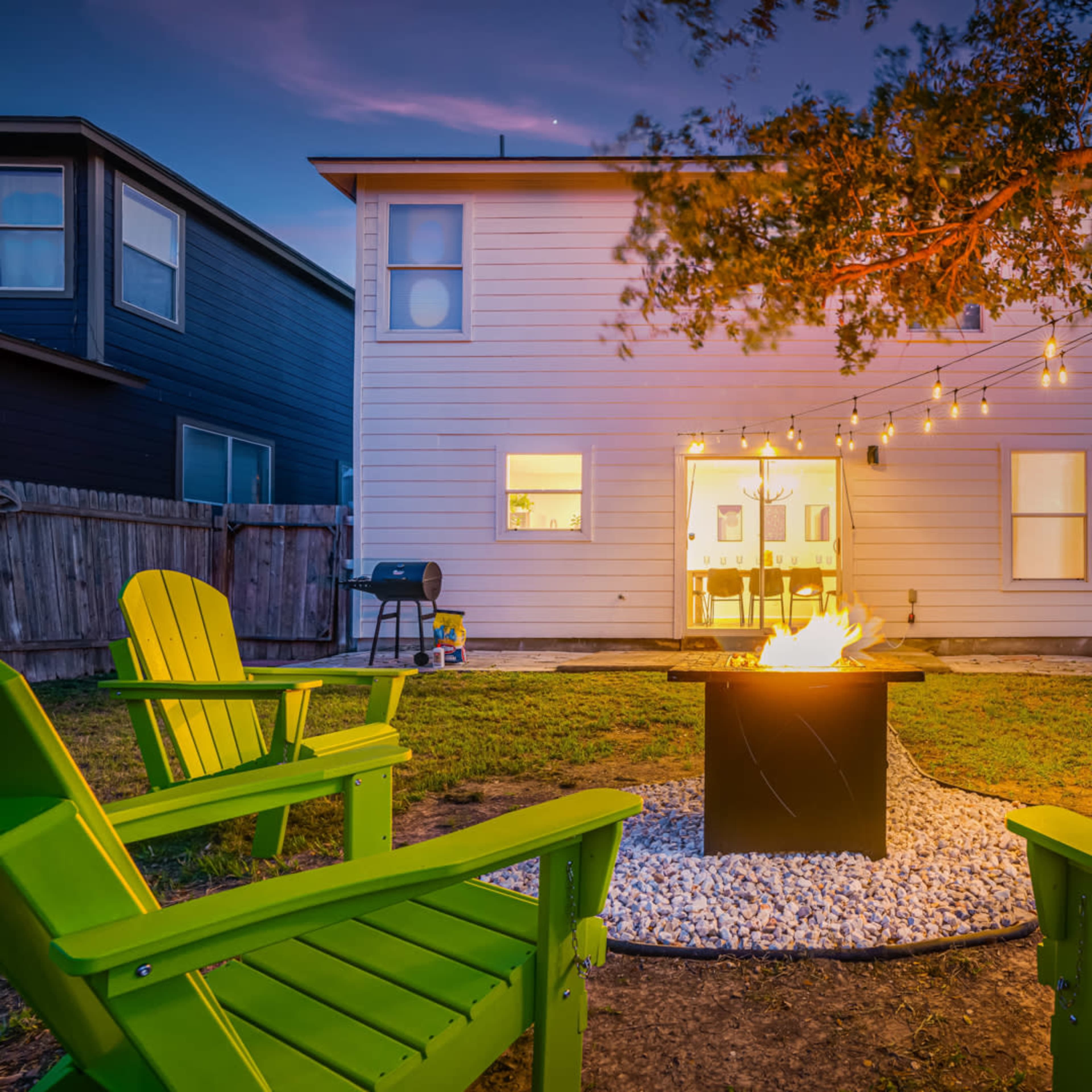 A backyard scene features a fire pit surrounded by four green chairs, with a house and string lights in the background.