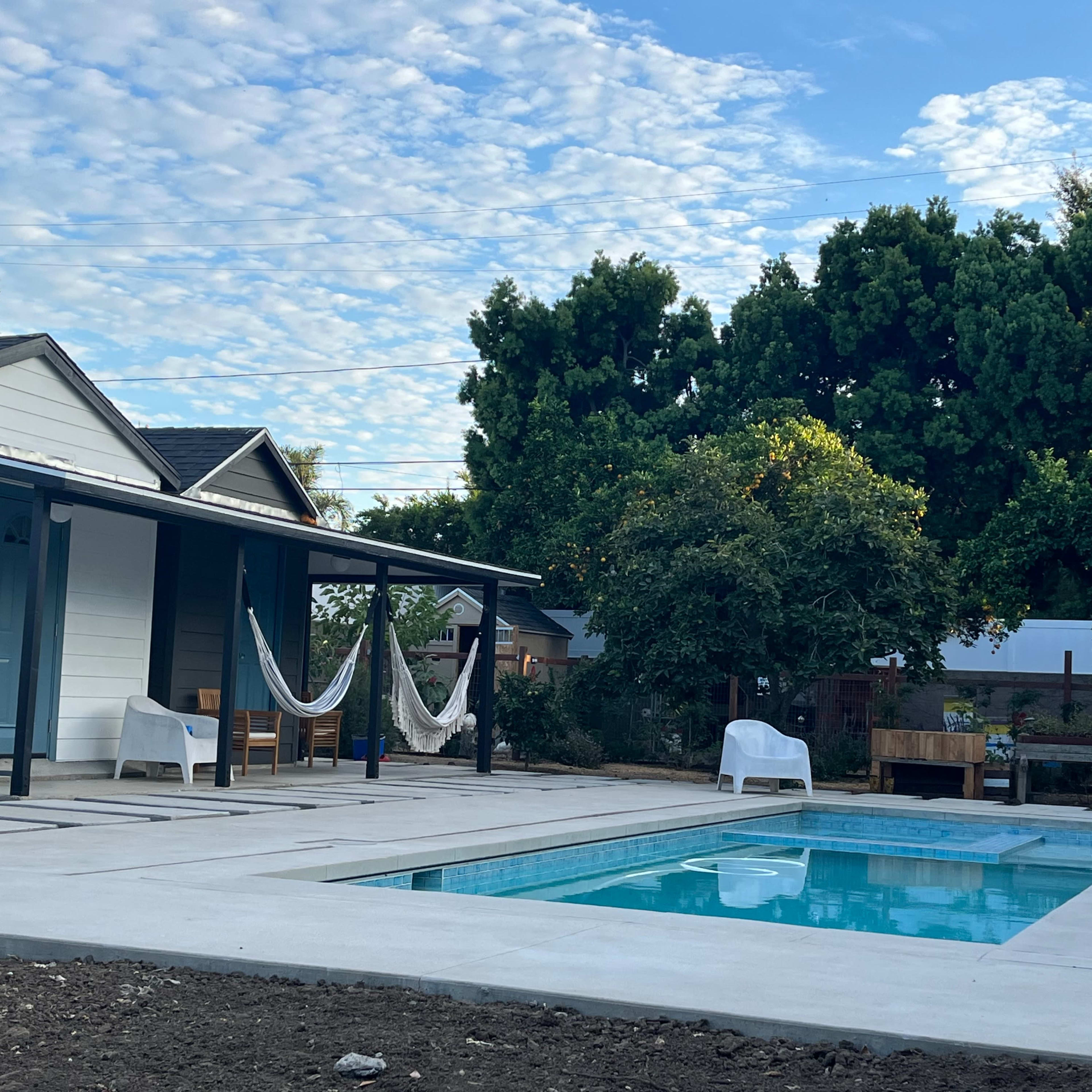 A rectangular swimming pool is situated between two houses, with hammocks hanging nearby and a garden area in the background.