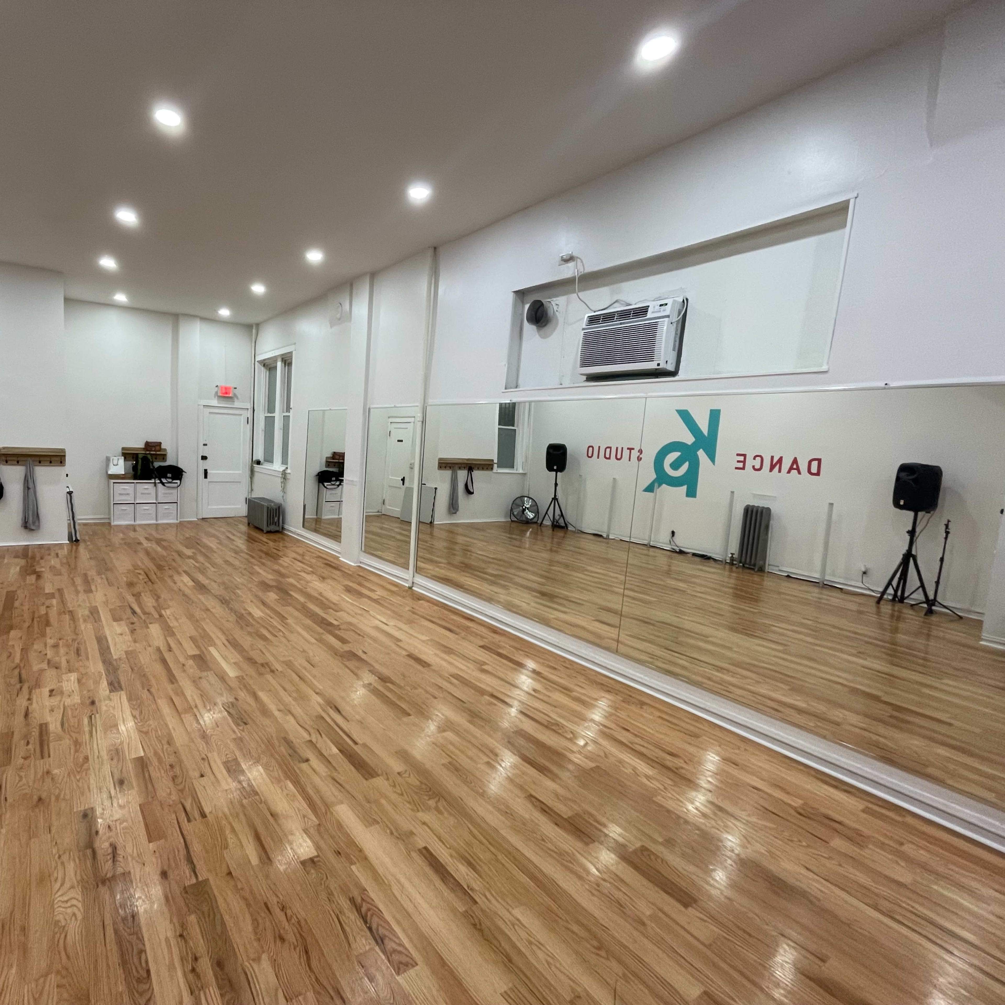 An empty dance studio featuring hardwood flooring, large mirrors on one wall, and a simple, brightly lit interior.