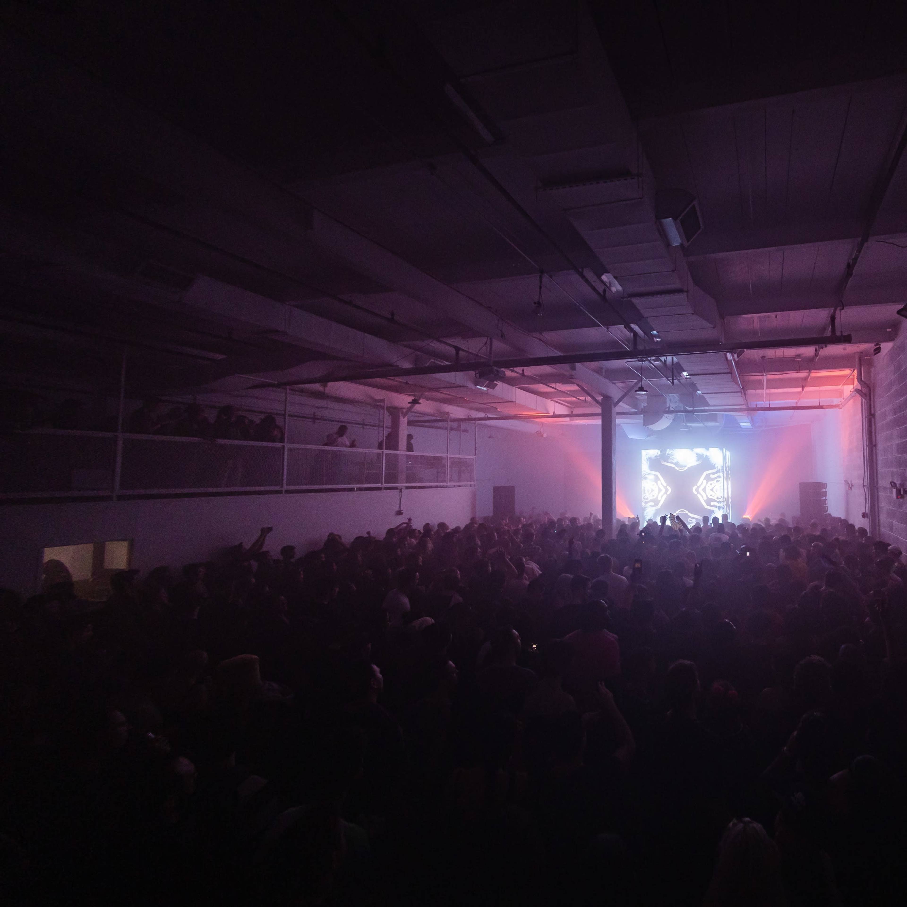 A large crowd fills a warehouse space, illuminated by colorful lighting and a projected visual display.