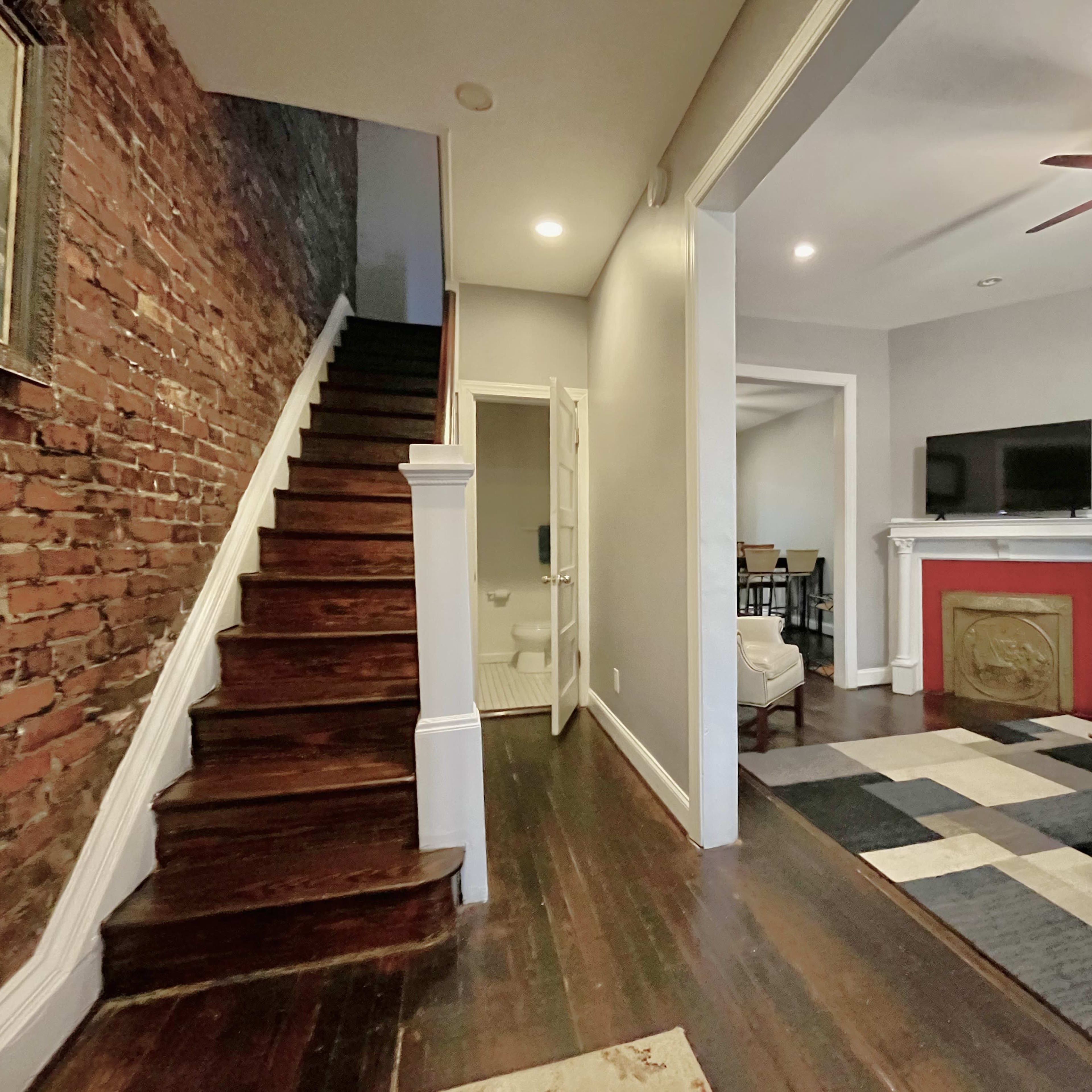 A hallway with exposed brick walls, a staircase leading upward, and a living area featuring a television on a mantel with a red surround.