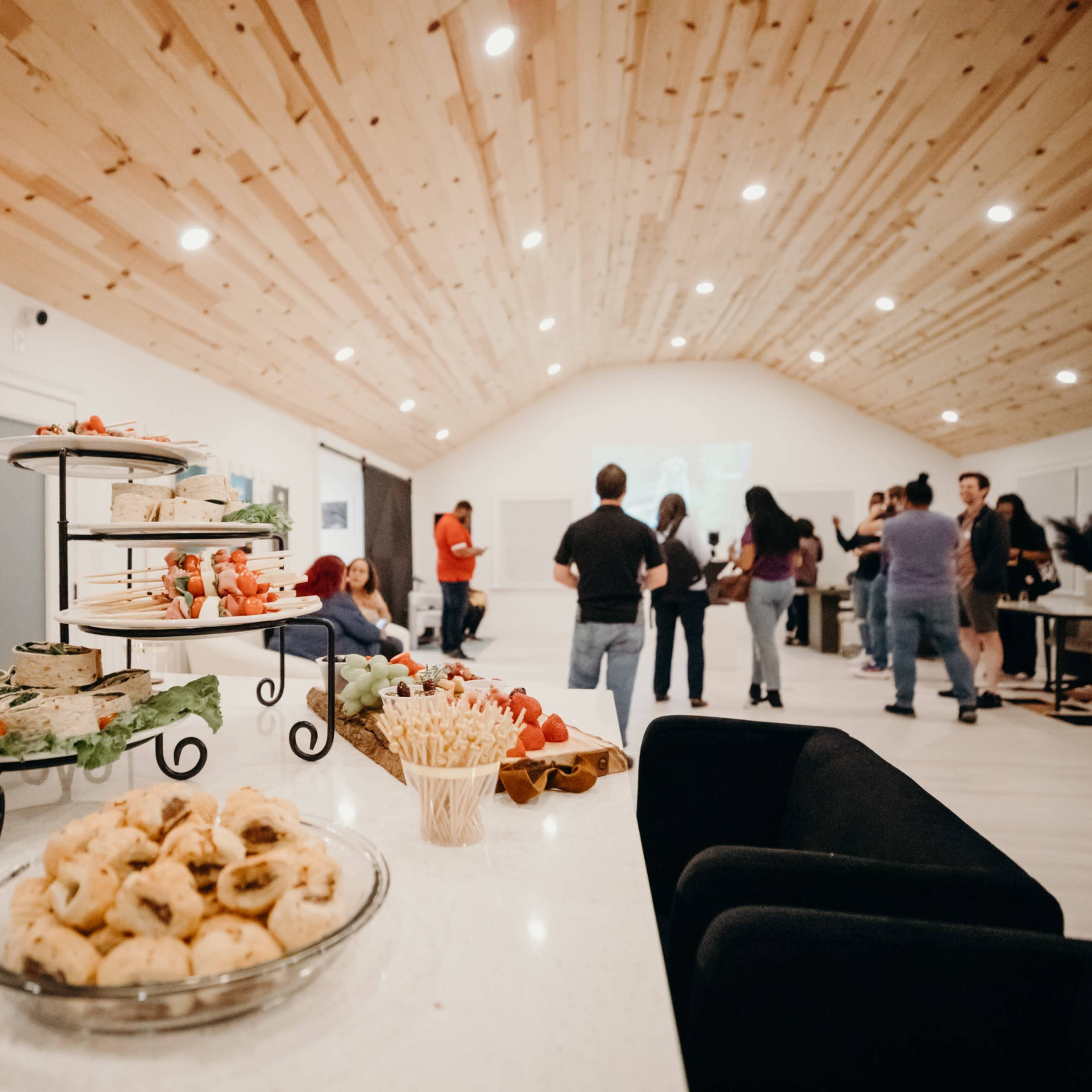 A gathering takes place in a bright room with a wooden ceiling, featuring a table with an array of food and drinks.